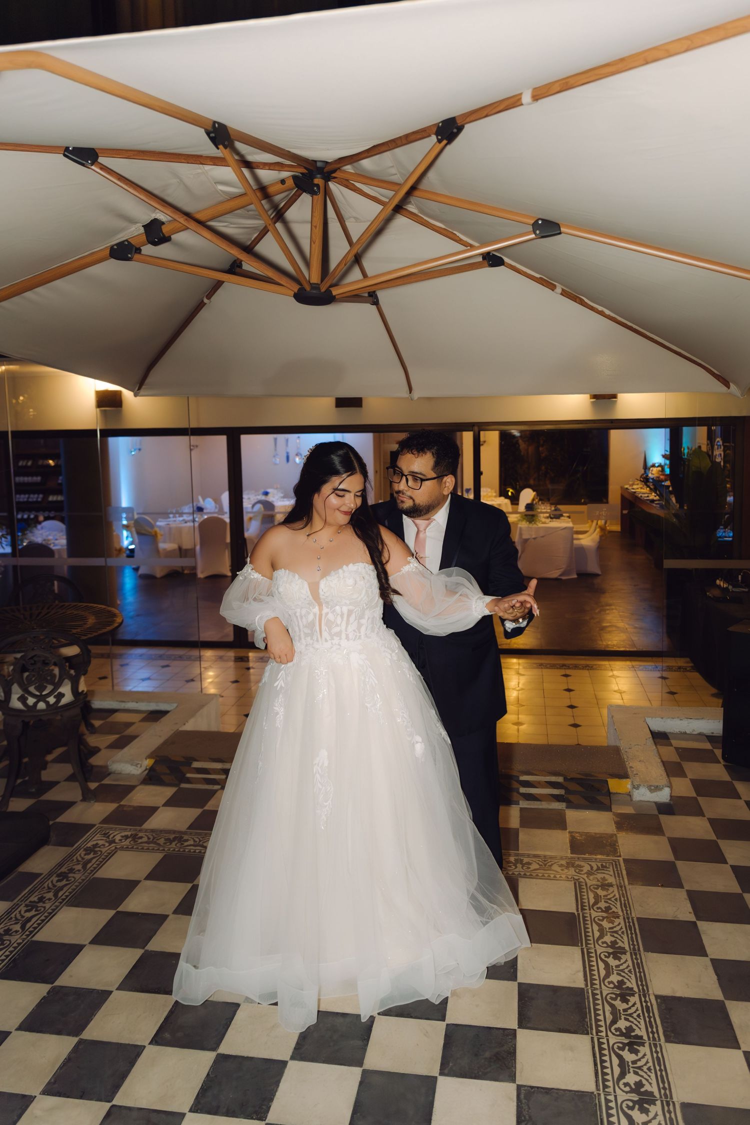 Bride in flowing white tulle gown and groom in navy suit sharing first dance under large patio umbrella.