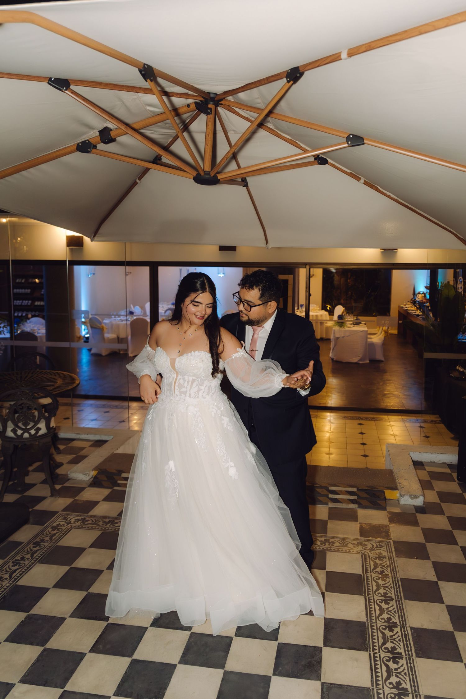 Newlyweds sharing intimate first dance on checkered tile floor beneath wooden umbrella at wedding venue.