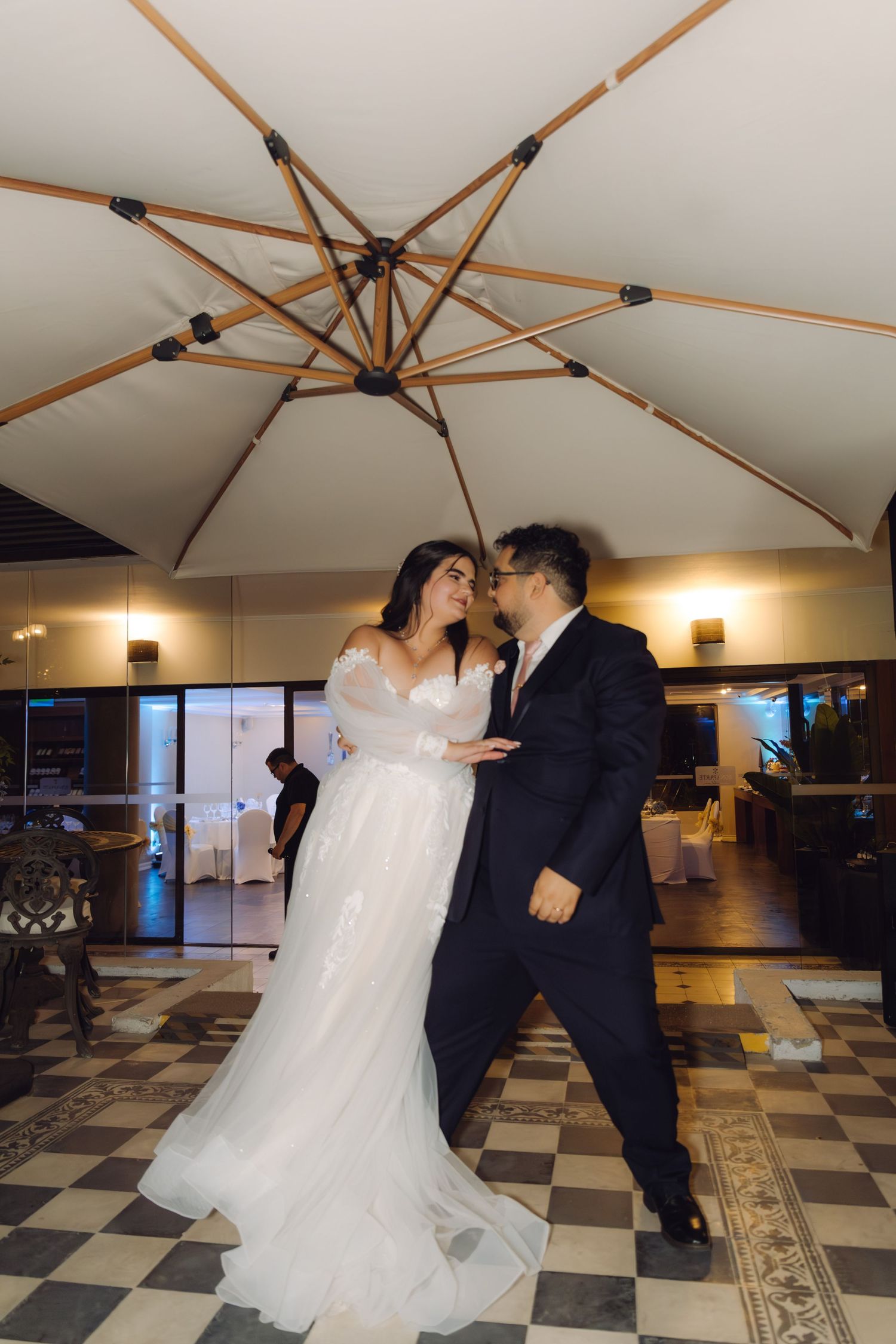 Bride and groom dancing closely together under wooden umbrella on elegant checkered ballroom floor.