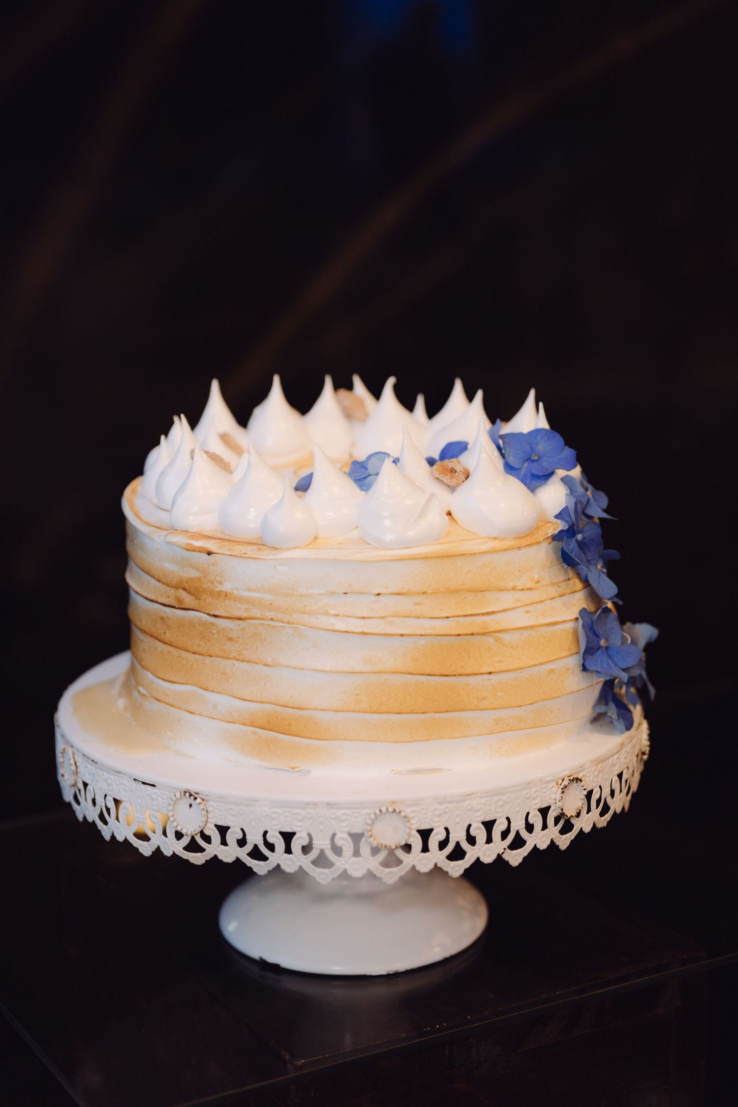 Elegant naked wedding cake topped with meringue kisses and blue hydrangea flowers on white cake stand.