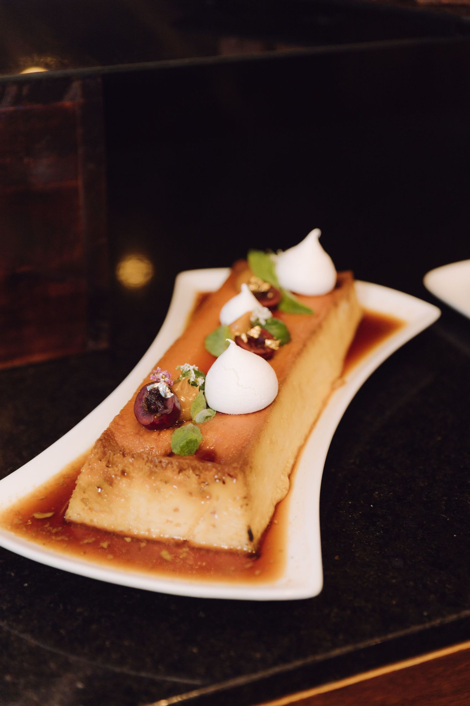 Elegant plated flan dessert with caramel sauce, cherries, meringue drops and micro herbs on white dish.