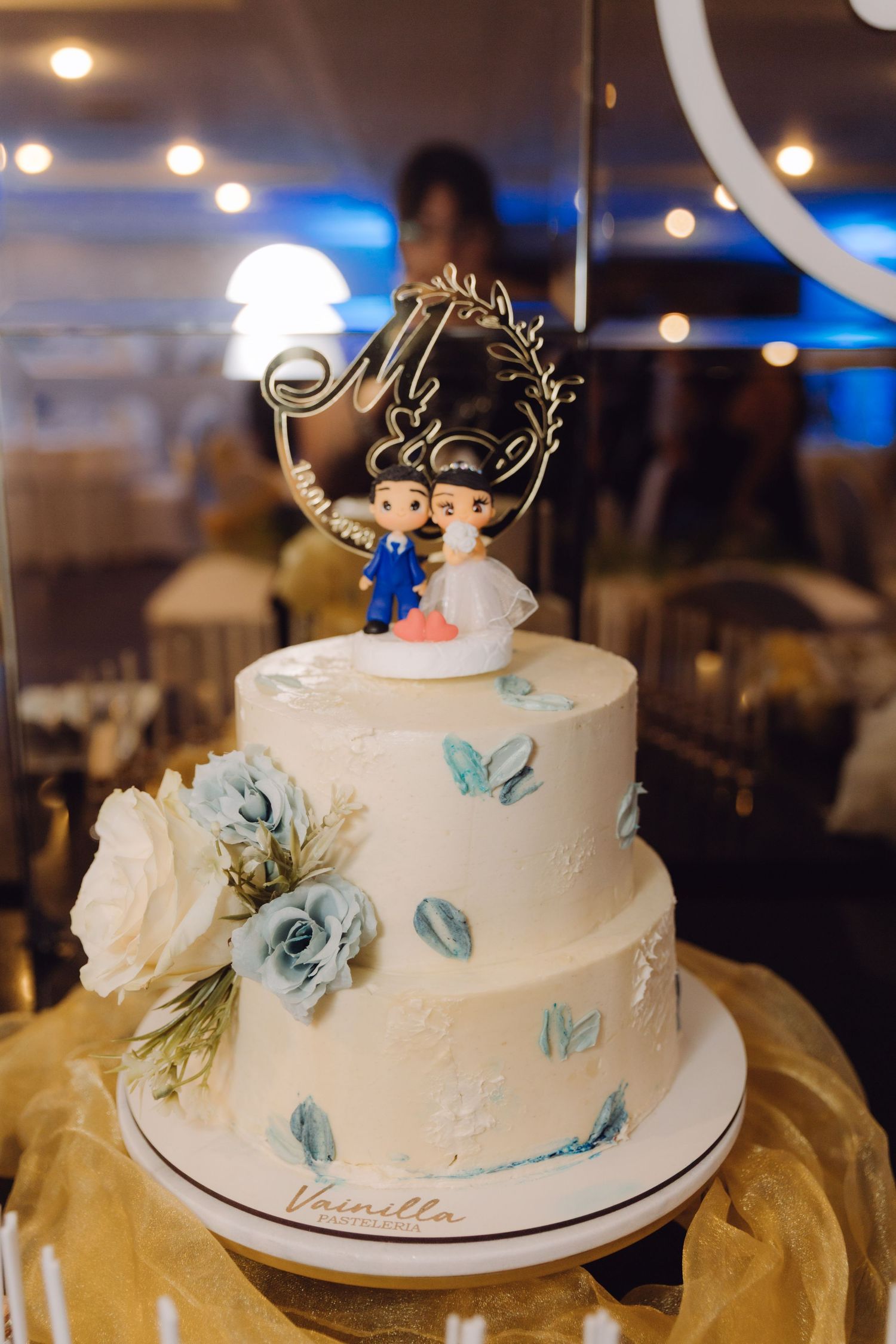 Two-tier white wedding cake with blue floral details and bride and groom figurine topper labeled Vanilla.
