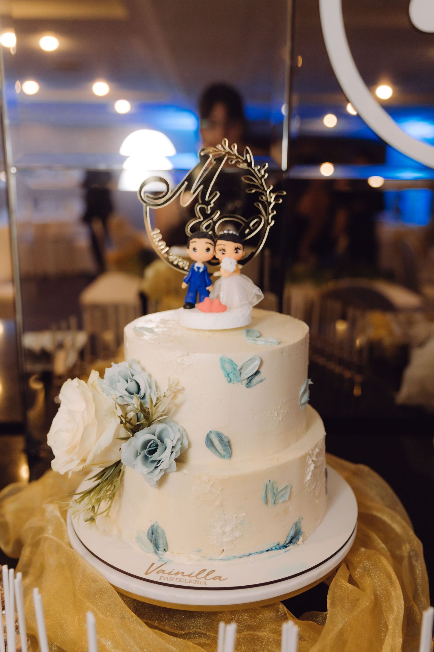 Elegant two-tier white wedding cake decorated with blue roses and couple figurines on top at a reception.