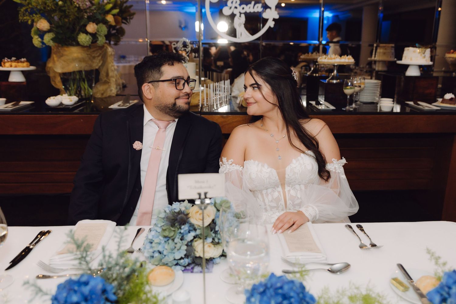 Bride and groom sitting together at a decorated reception table with blue floral centerpieces indoors.
