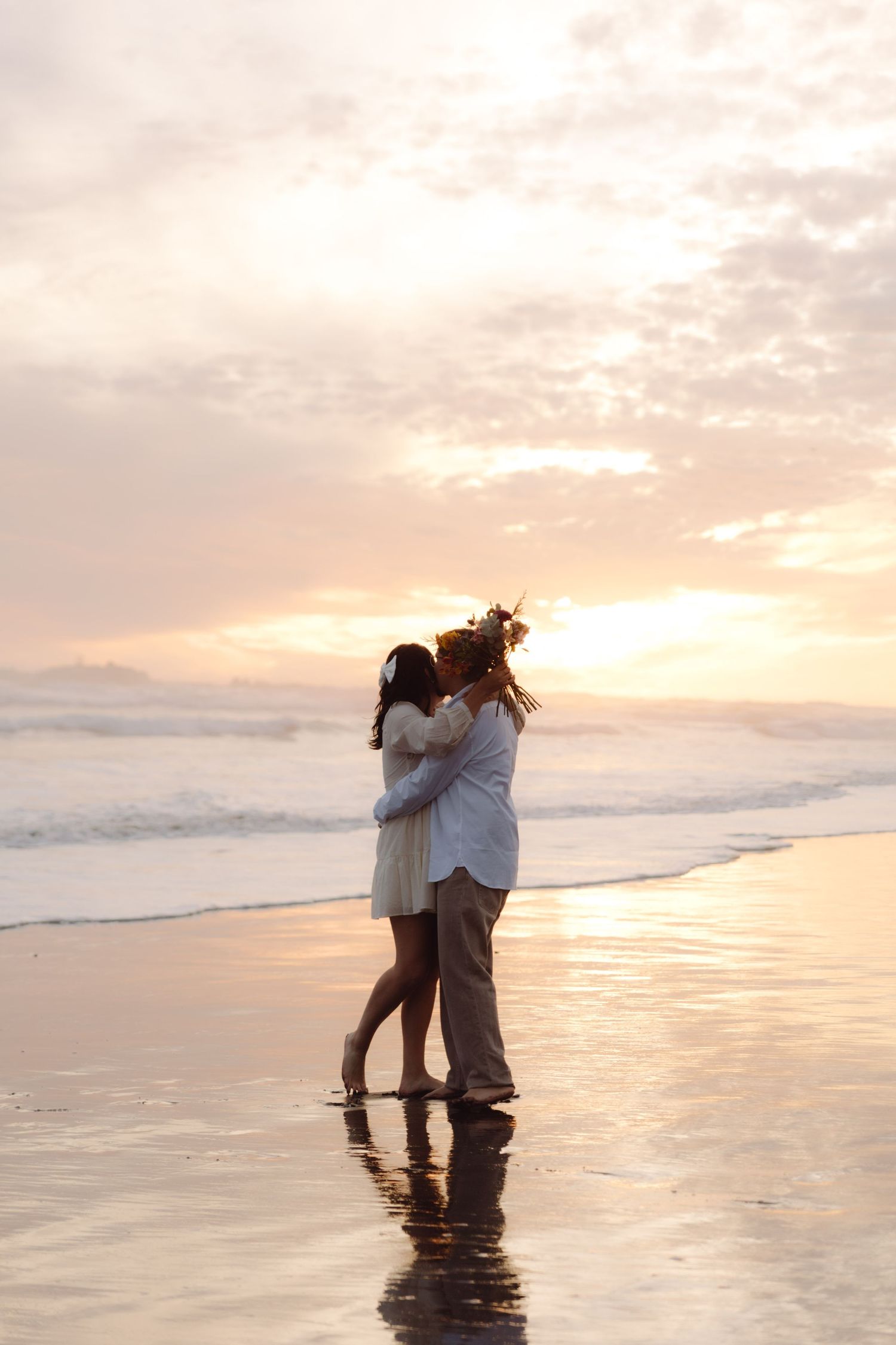 Romantic couple kissing at the shoreline during a golden sunset beach elopement with a colorful bouquet.