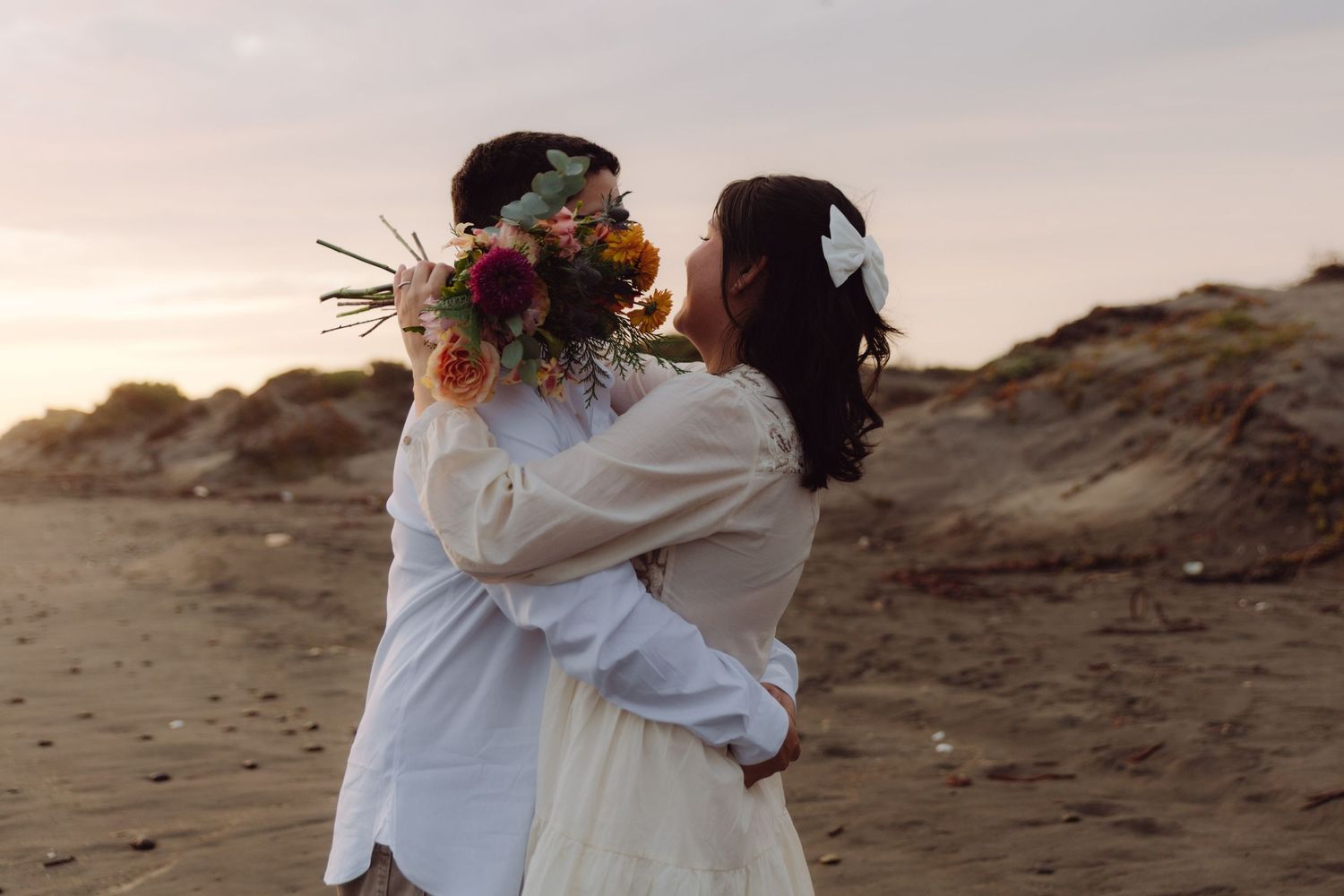 Couple sharing a kiss on a sandy beach at dusk, bride holding a vibrant floral bouquet with a white bow.