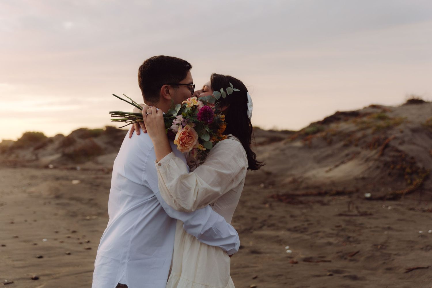 Couple embracing and kissing on a beach at sunset, bride holding a colorful bouquet in a white dress.