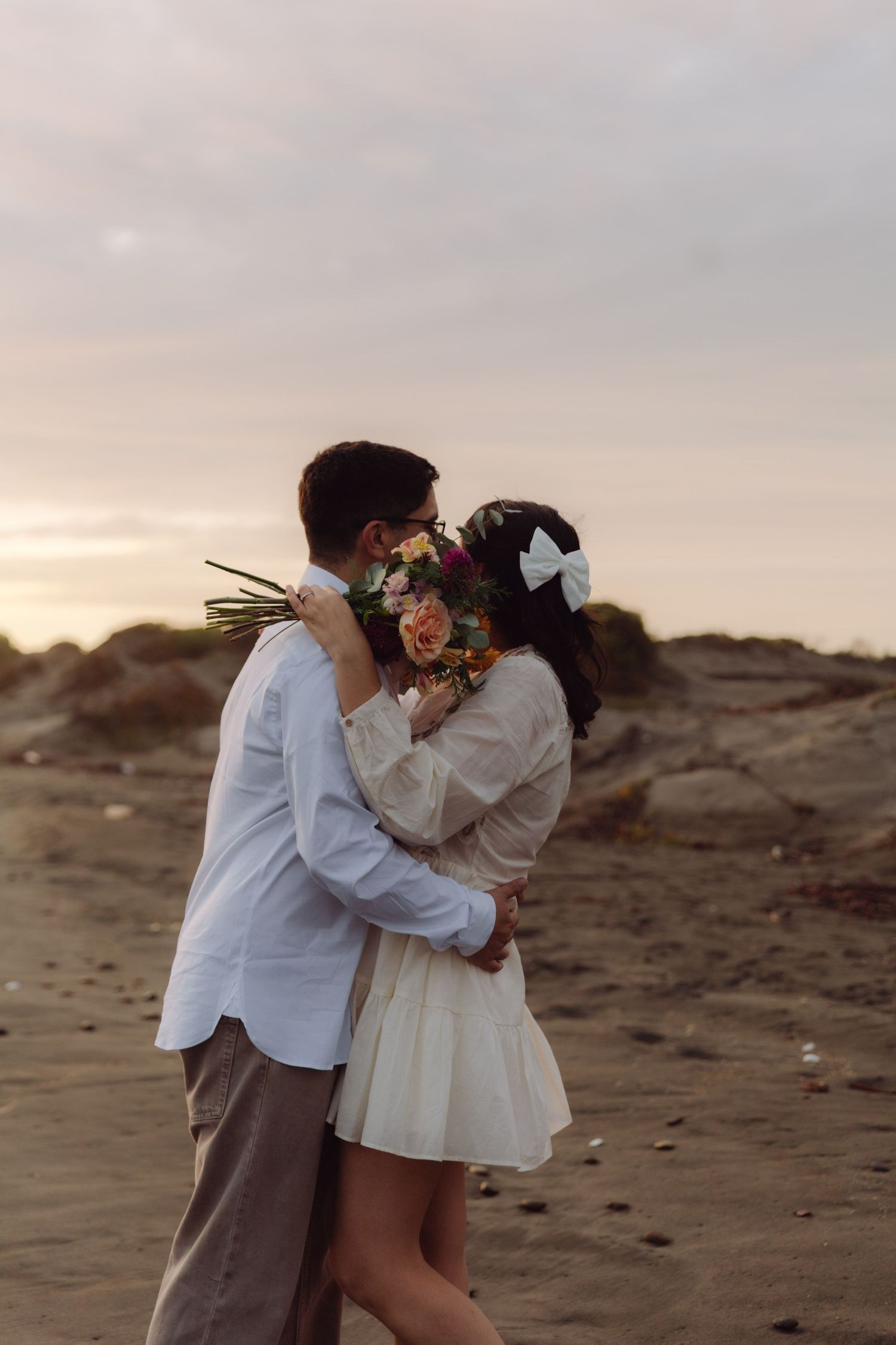 Couple kissing on a sandy beach at sunset, bride in white dress holding a bouquet with a white hair bow.