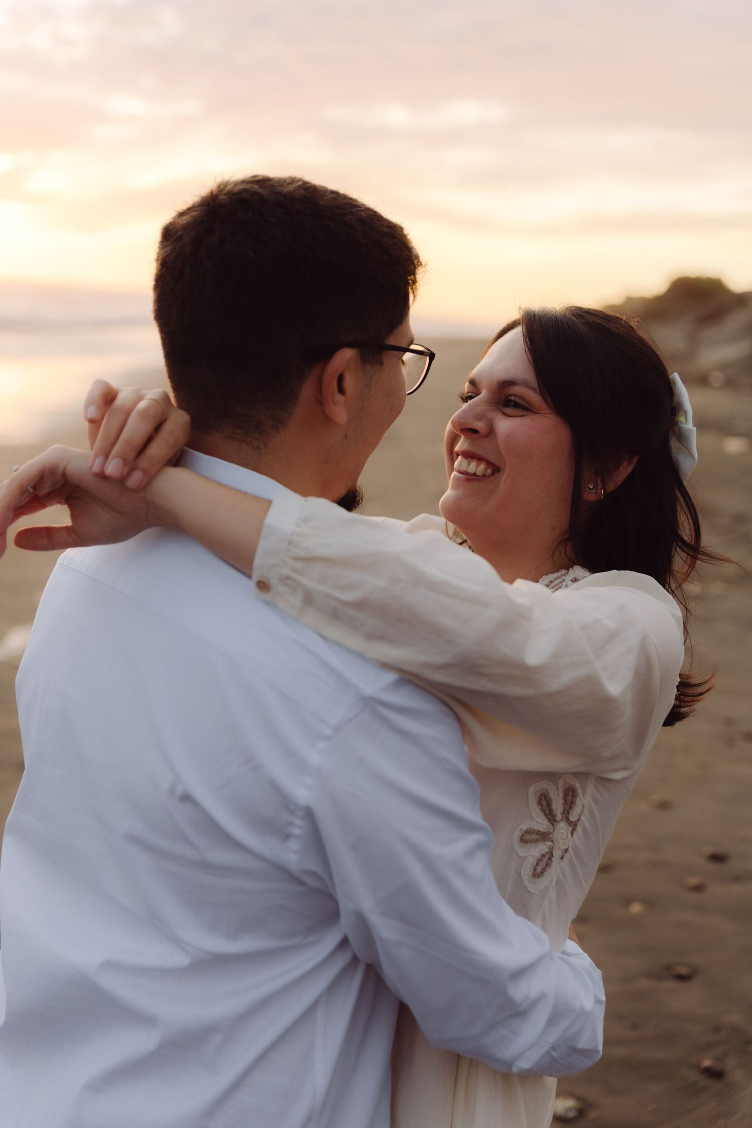 Bride laughing joyfully while embracing her partner on a beach during a romantic sunset elopement session.