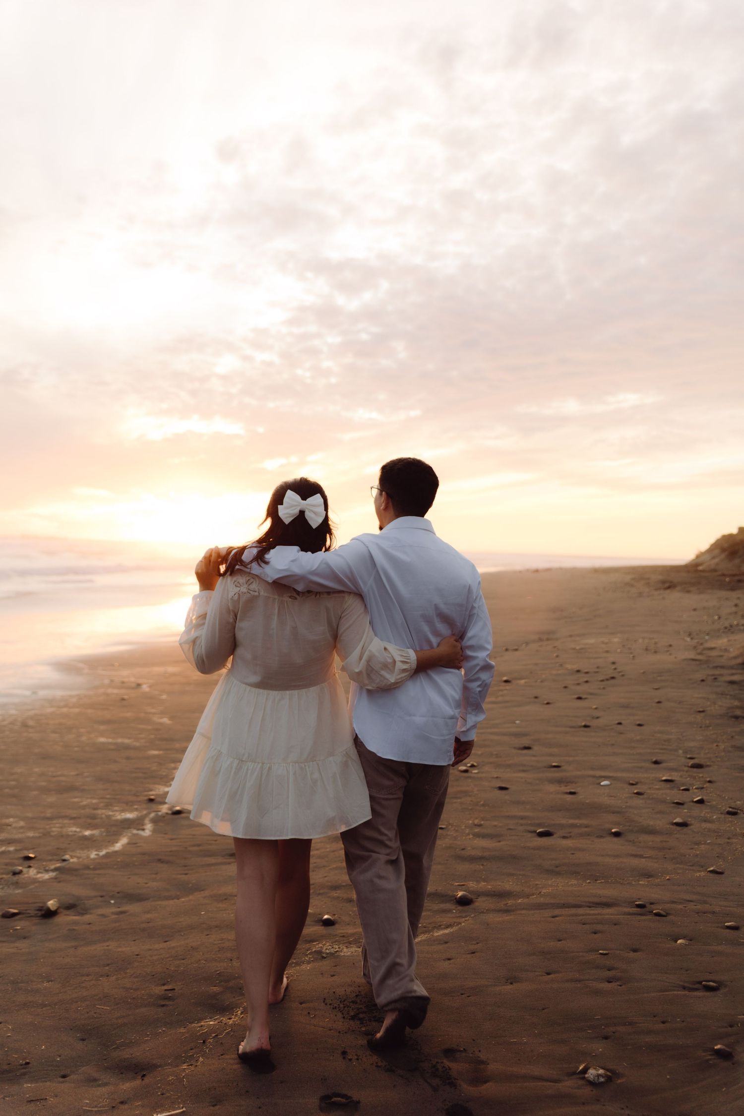 Couple walking arm in arm along a sandy beach toward a pastel sunset, viewed from behind.