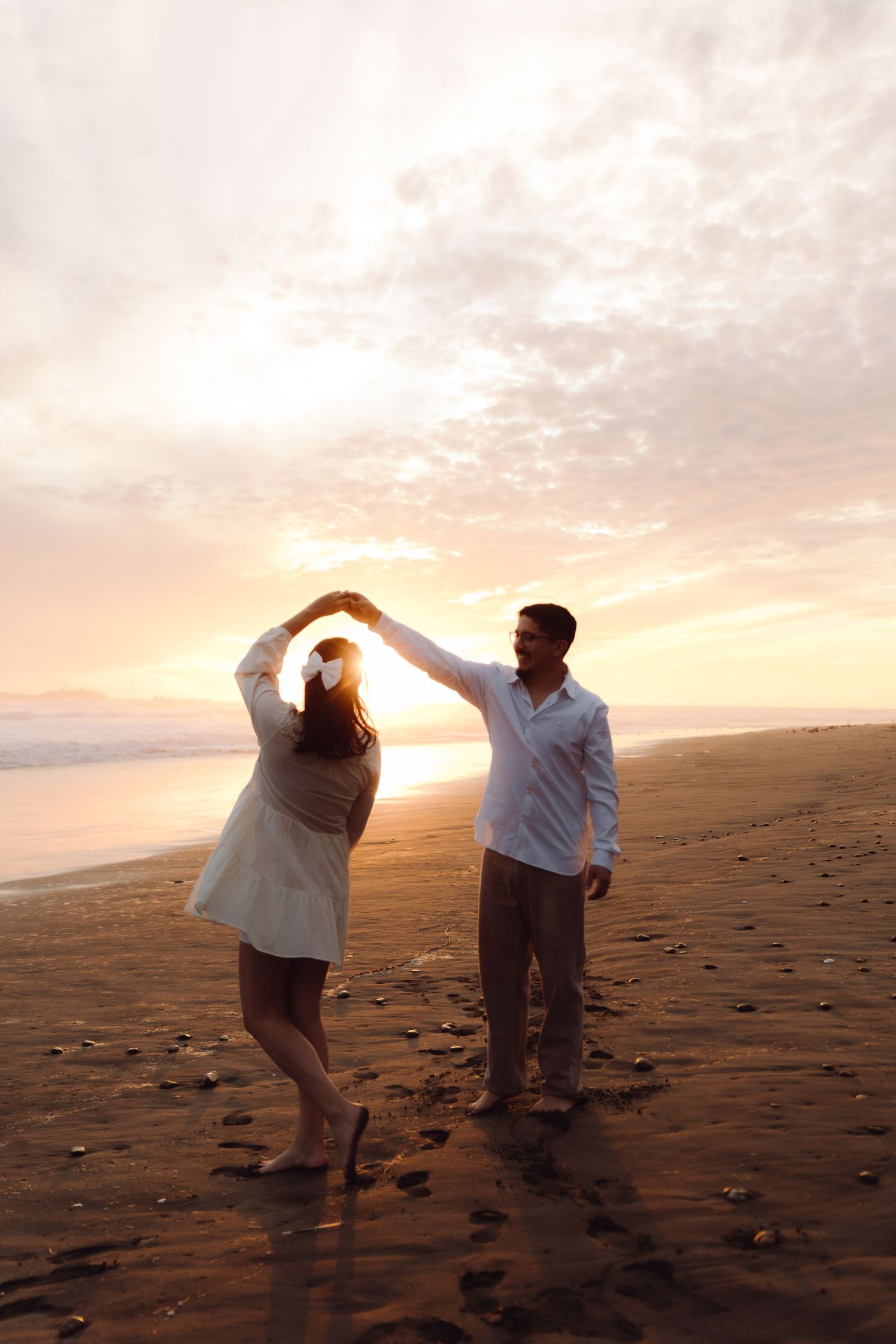 Couple dancing joyfully on a beach at sunset, man spinning woman under raised hands in golden light.
