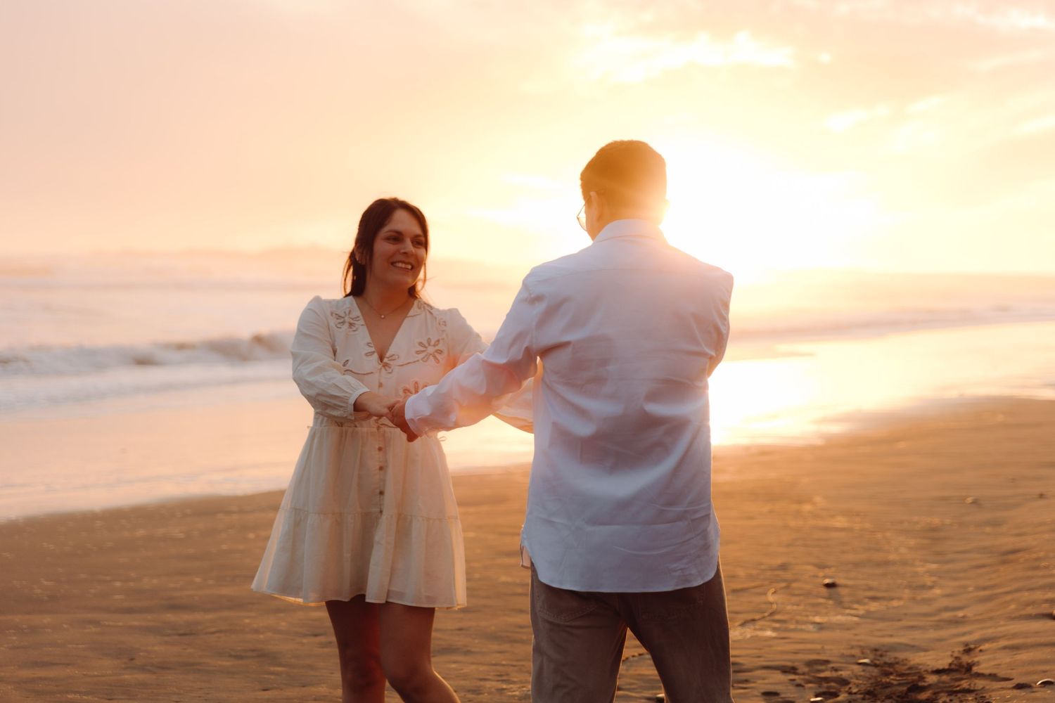 Couple holding hands on a sandy beach at golden sunset, woman in white dress smiling at man in light blue shirt.