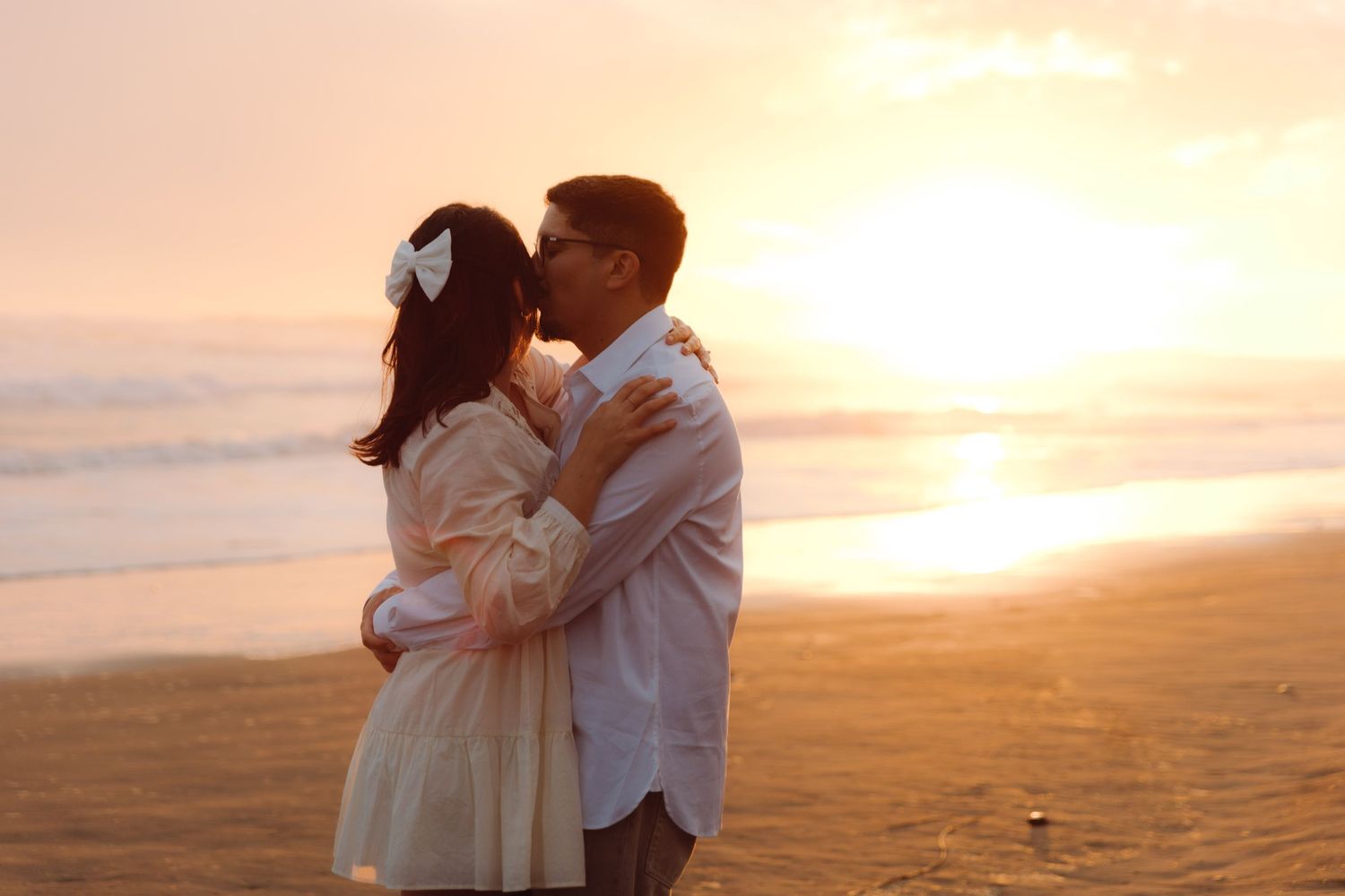 Romantic couple sharing a kiss on the beach during a warm golden sunset, woman wearing a white hair bow.