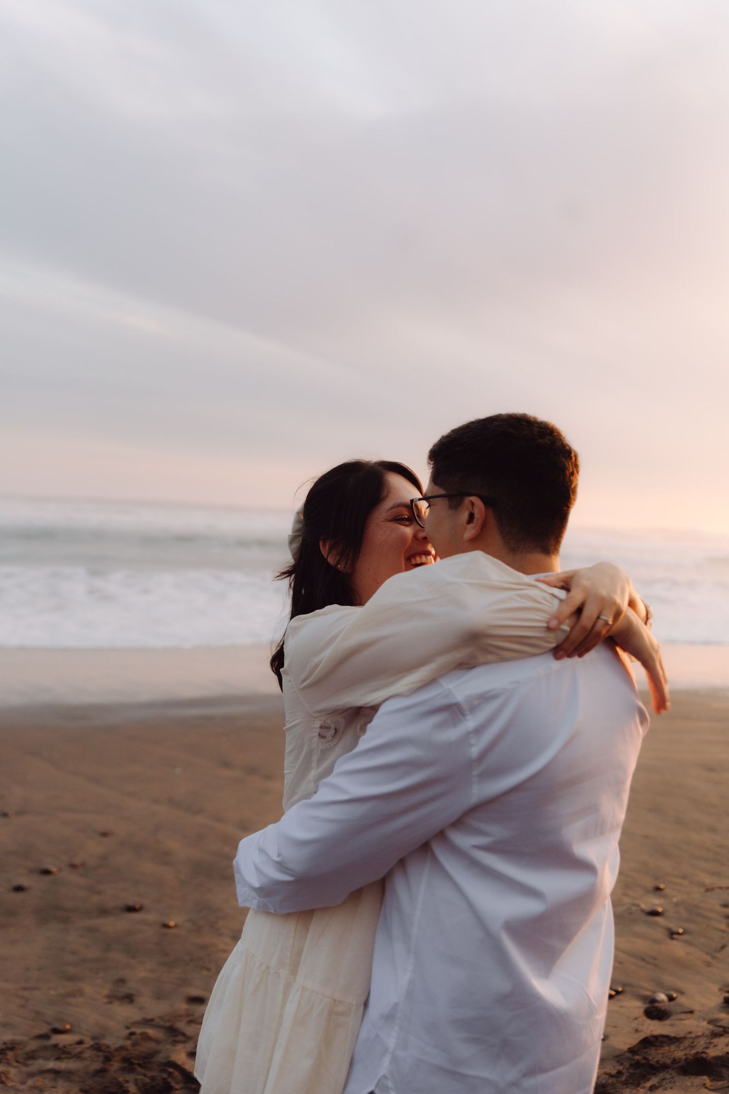 Couple embracing tenderly on a beach at dusk, woman in white dress hugging man in white shirt by ocean waves.