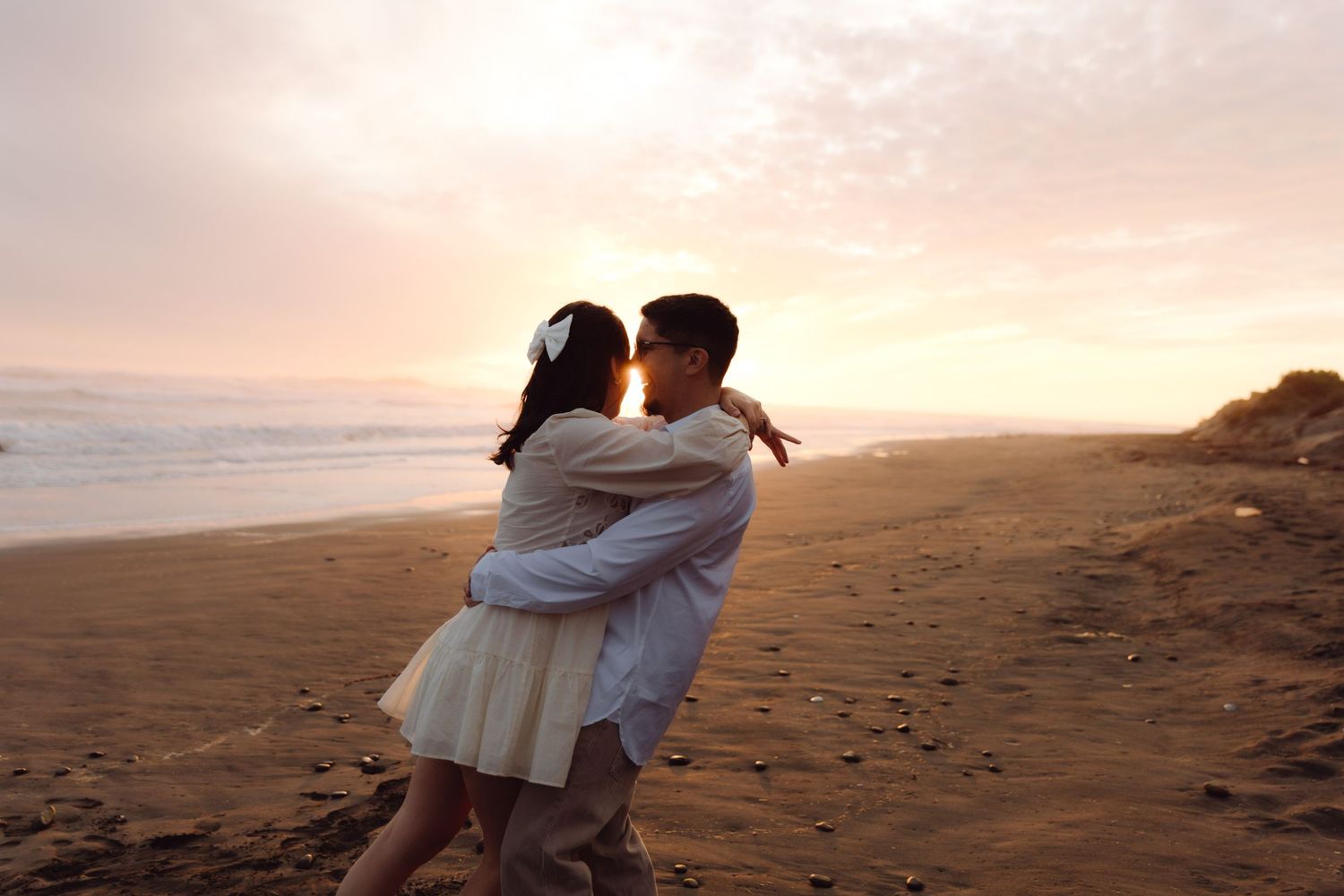 Couple dancing and embracing on a sandy beach at sunset, woman in white mini dress and man in white shirt.