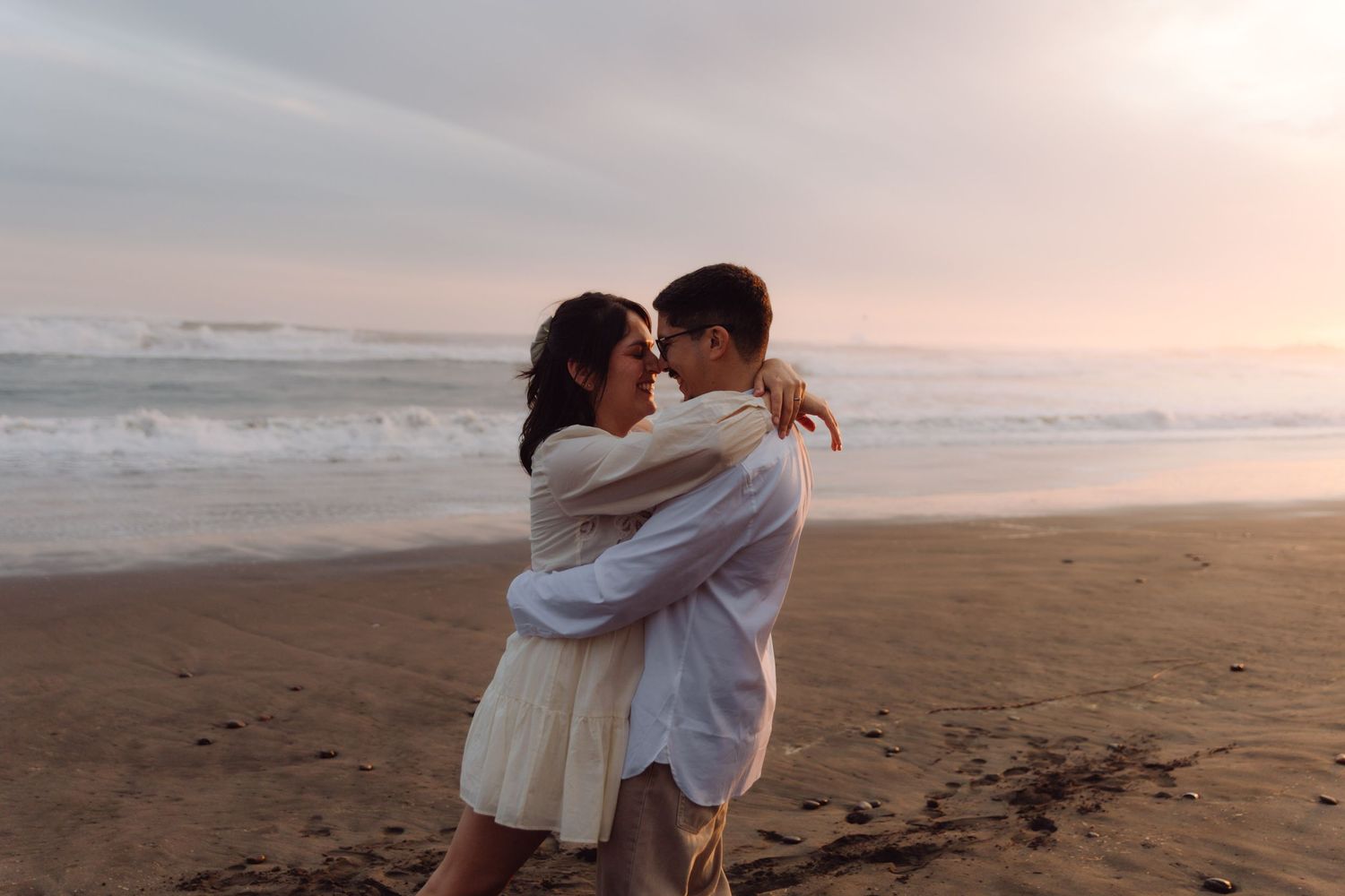 Loving couple holding each other close on a beach at pastel sunset, smiling and sharing an intimate moment.