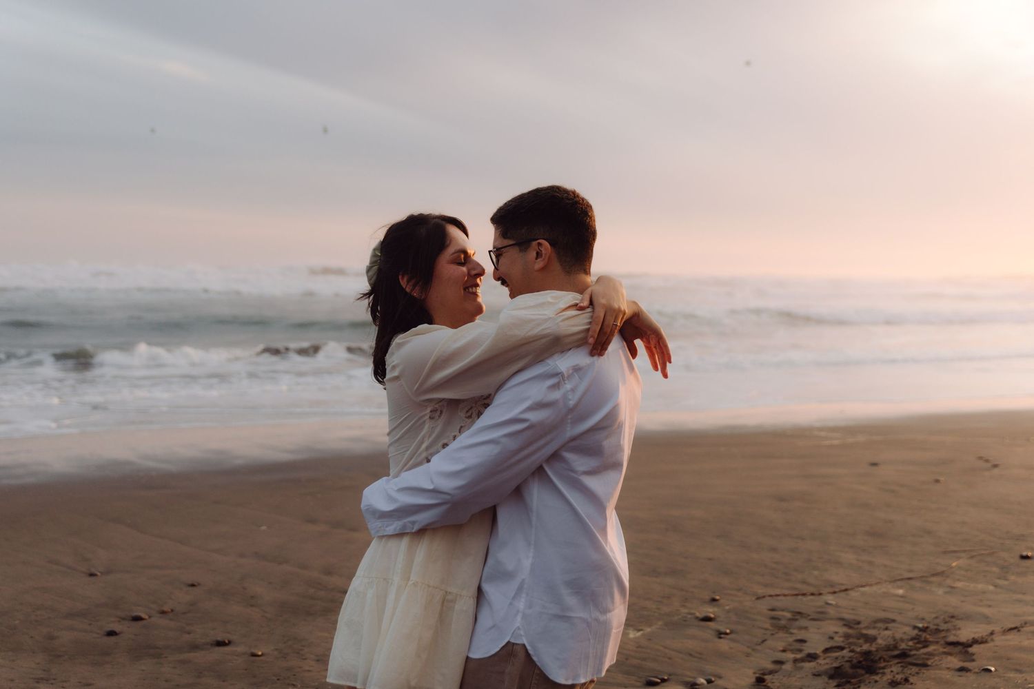 Couple sharing a romantic embrace on a sandy beach at sunset, nearly kissing with ocean waves in background.