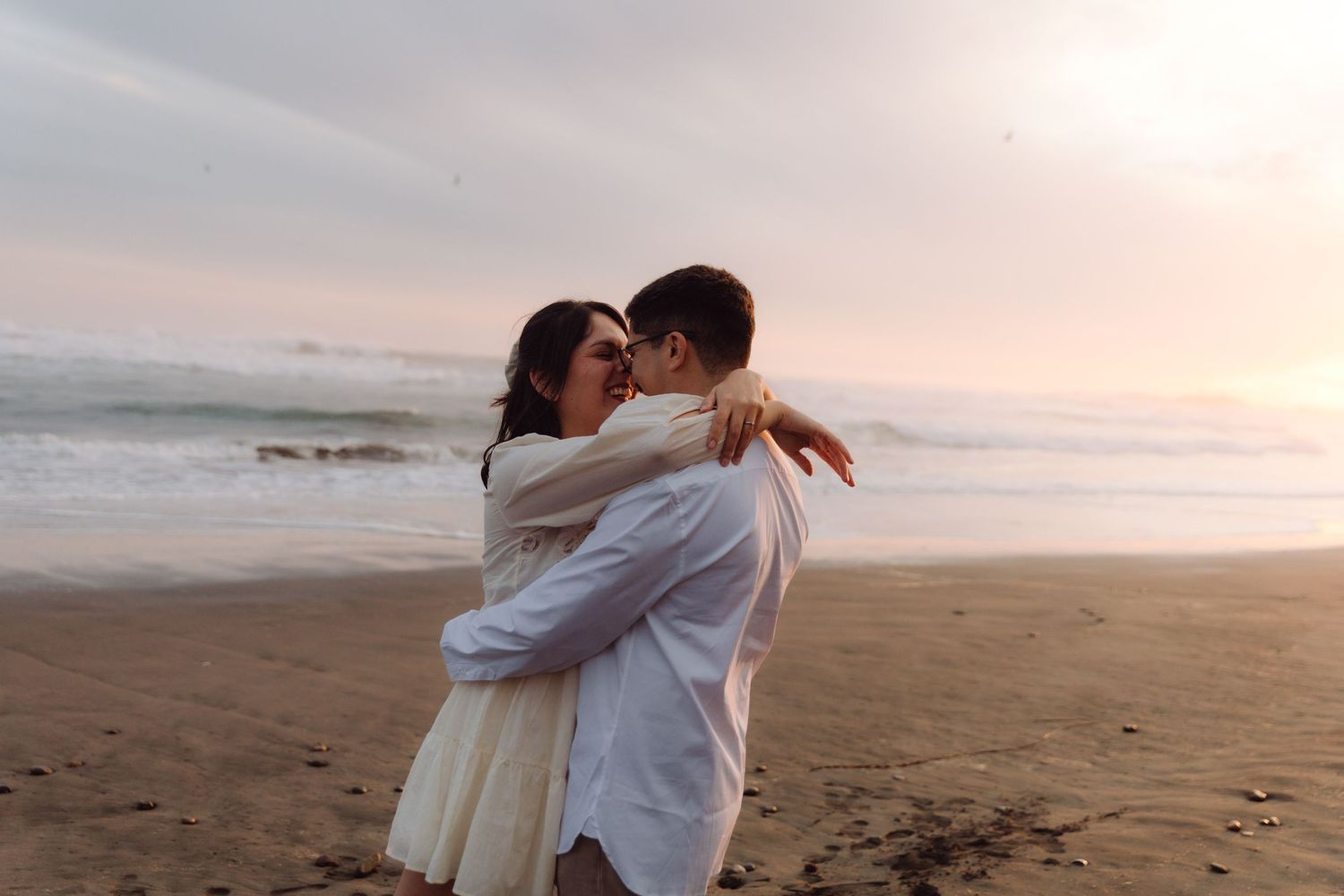 Couple hugging affectionately on a beach at dusk, woman in white dress and man in light blue shirt by the sea.