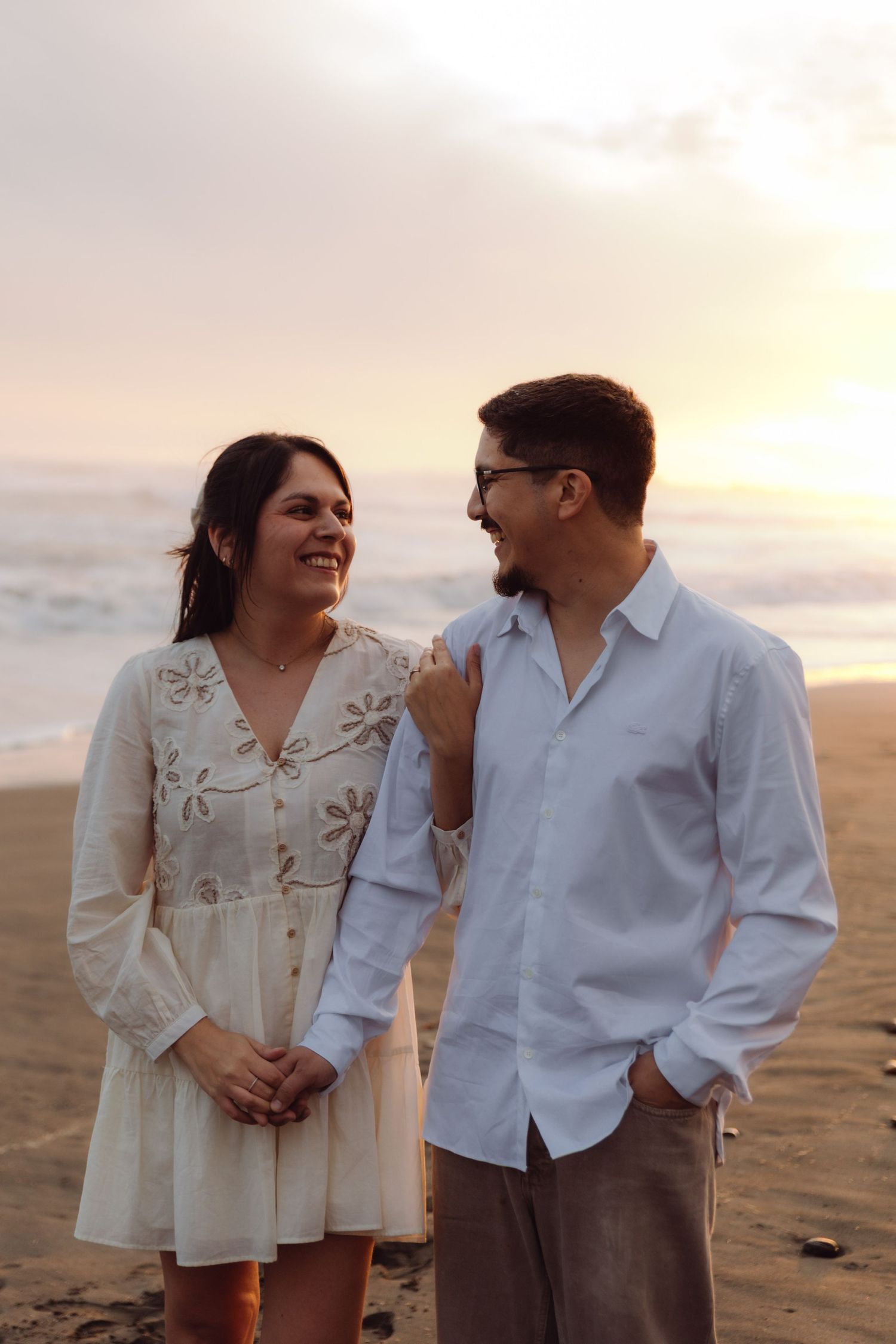 Happy couple laughing together on a beach at sunset, woman in embroidered white dress and man in blue shirt.