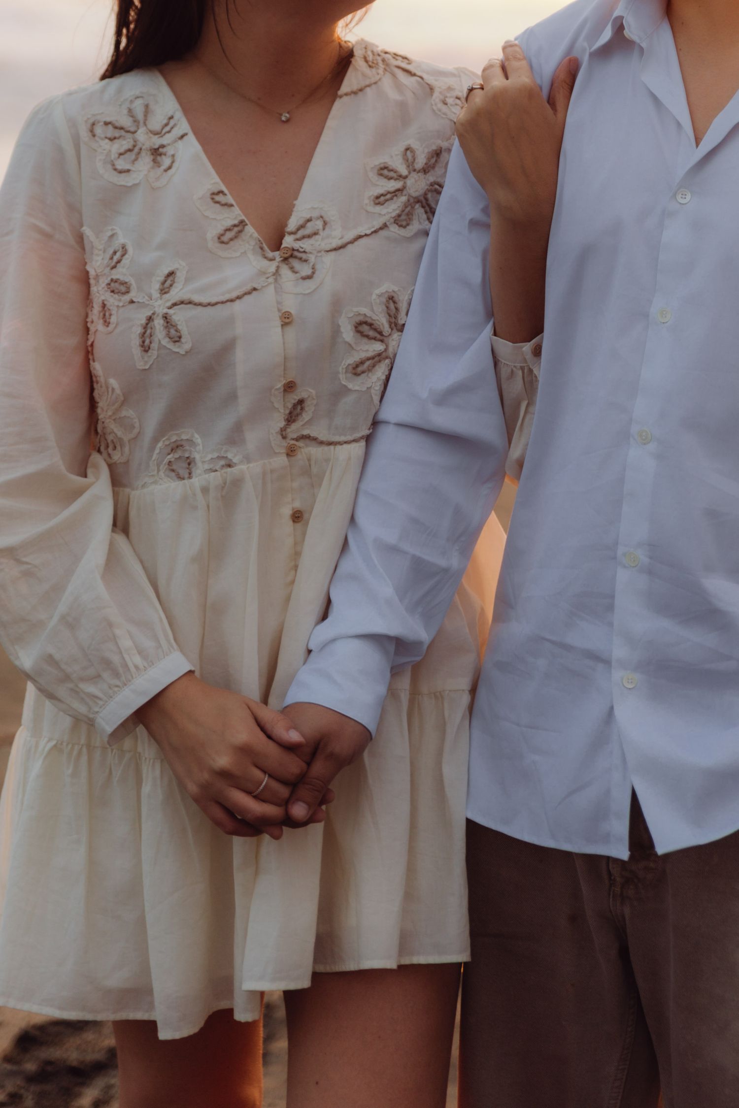 Close-up of couple holding hands on beach, woman in elegant embroidered white floral dress with sheer sleeves.