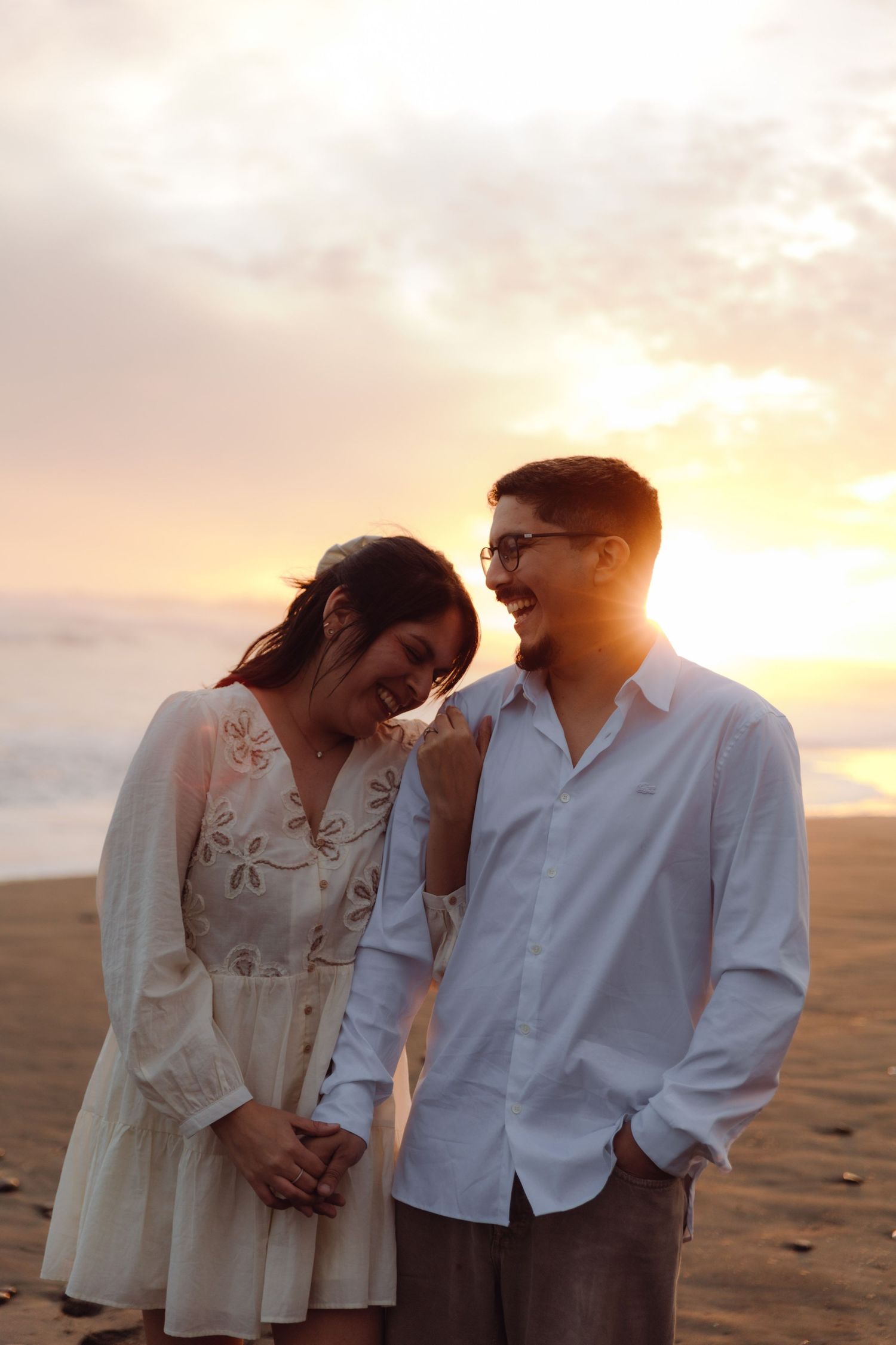 Joyful couple laughing and holding hands on a beach at golden sunset, woman in white embroidered dress.