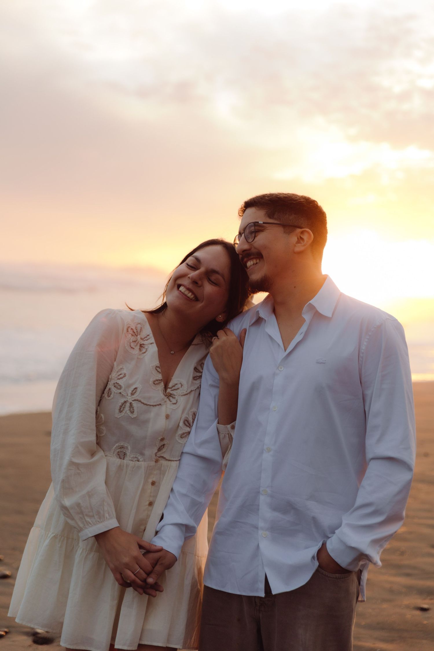 Couple laughing and holding hands on beach at golden sunset, woman in white dress leaning on mans shoulder.