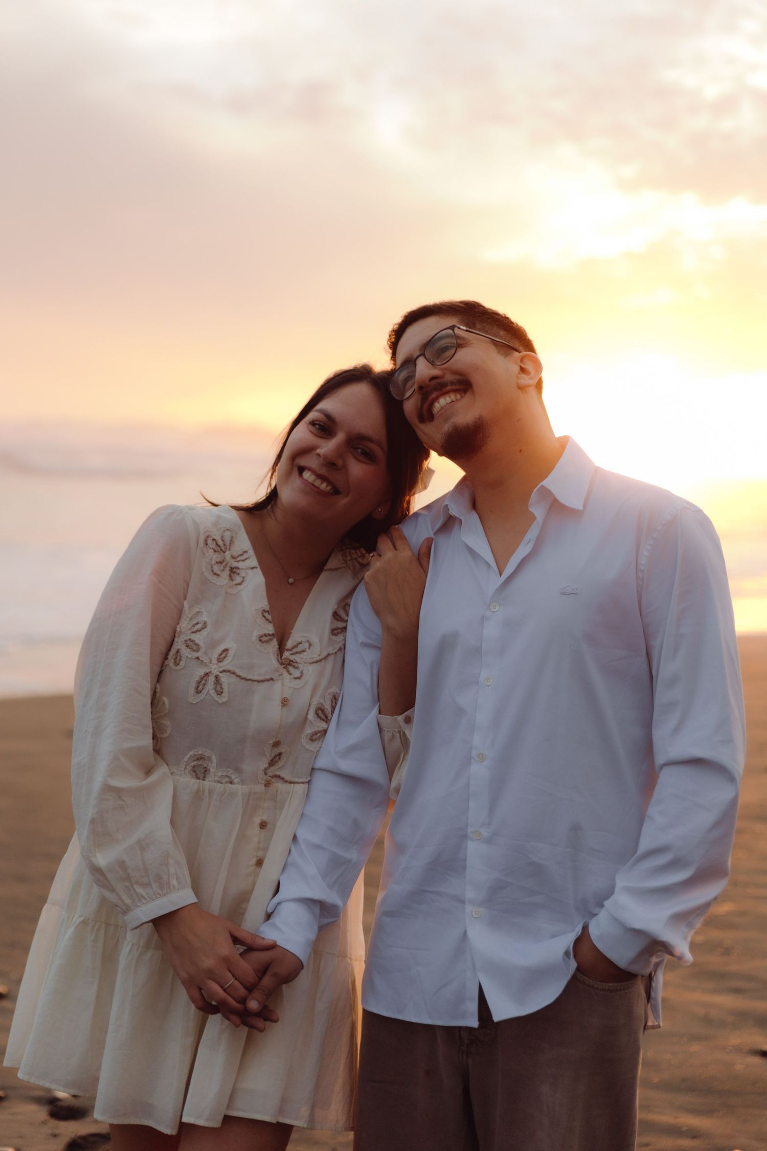 Happy couple smiling and holding hands on sandy beach during vibrant orange and pink sunset.