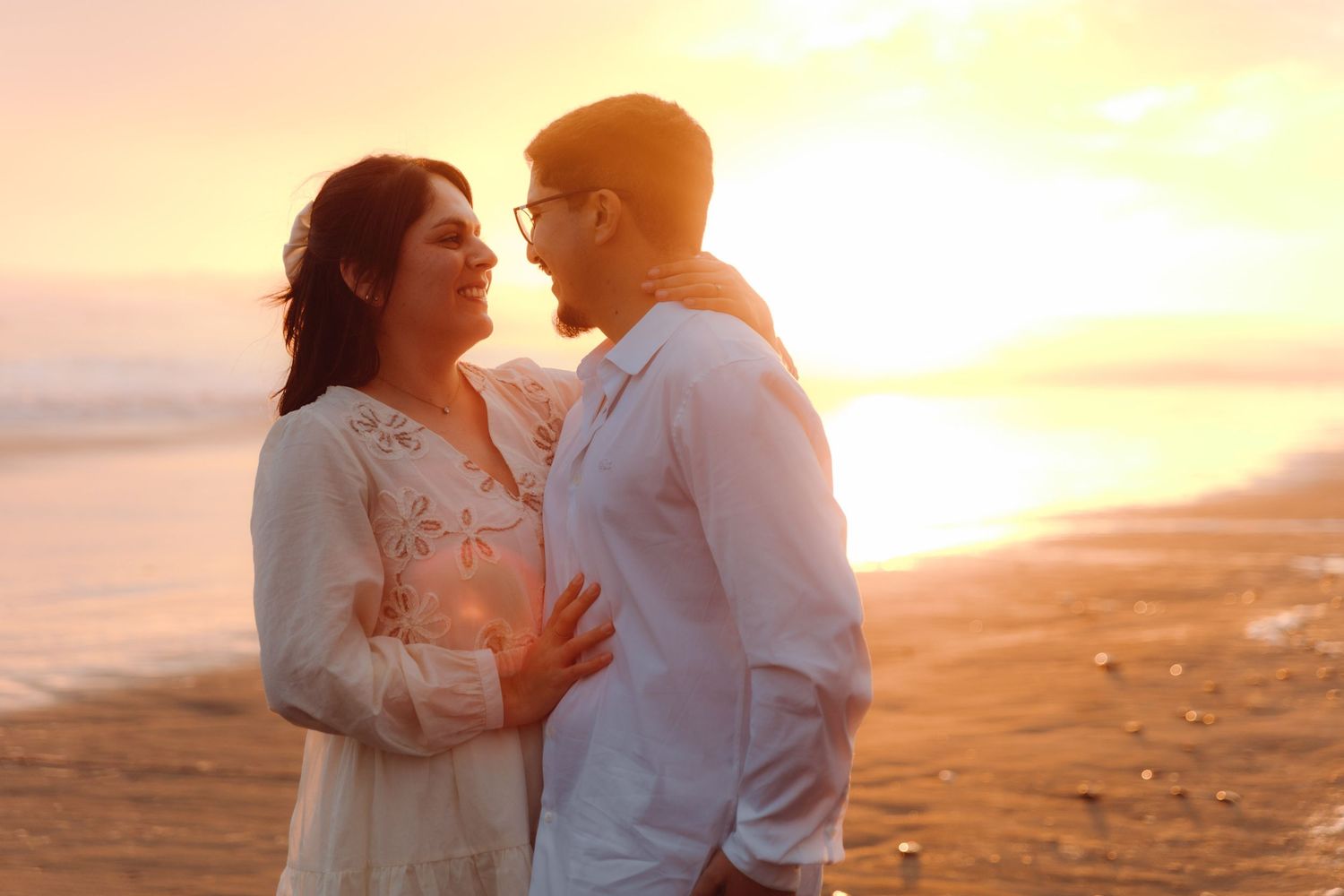 Romantic couple embracing face to face on beach with glowing golden sunset behind them.