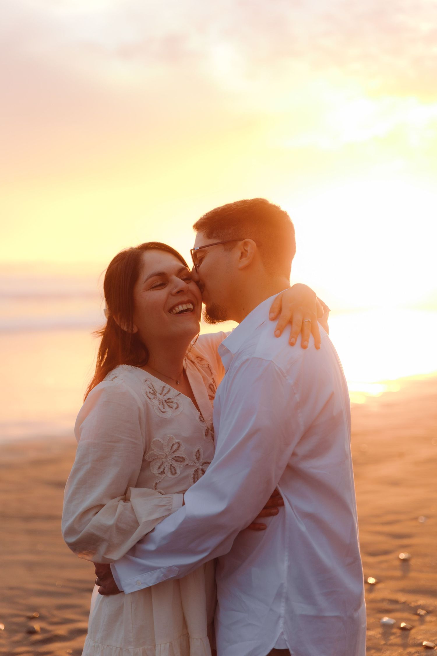 Man kissing woman on cheek as she laughs joyfully on beach during warm sunset.