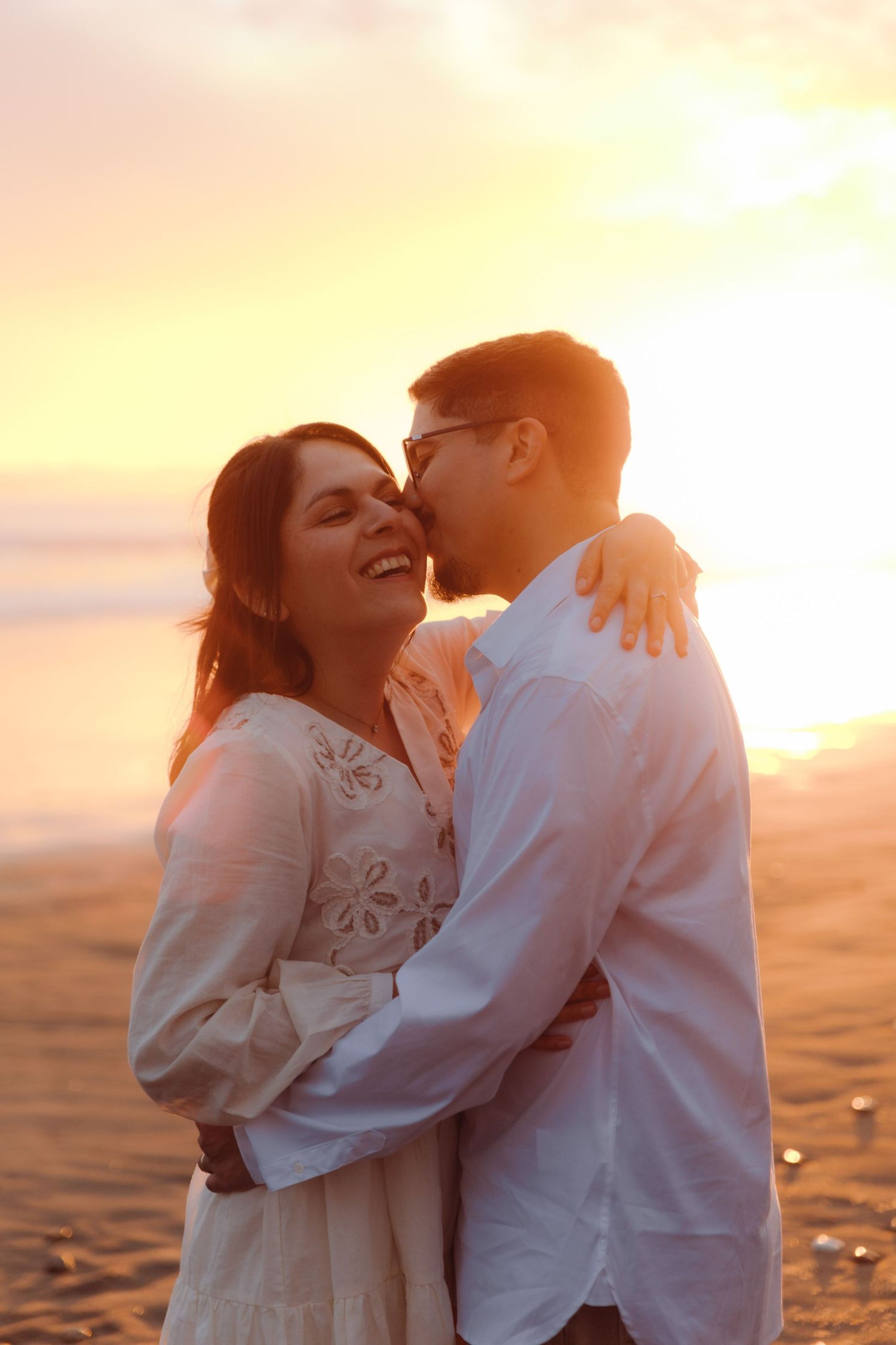 Couple hugging and laughing together on beach with bright golden sunset in background.