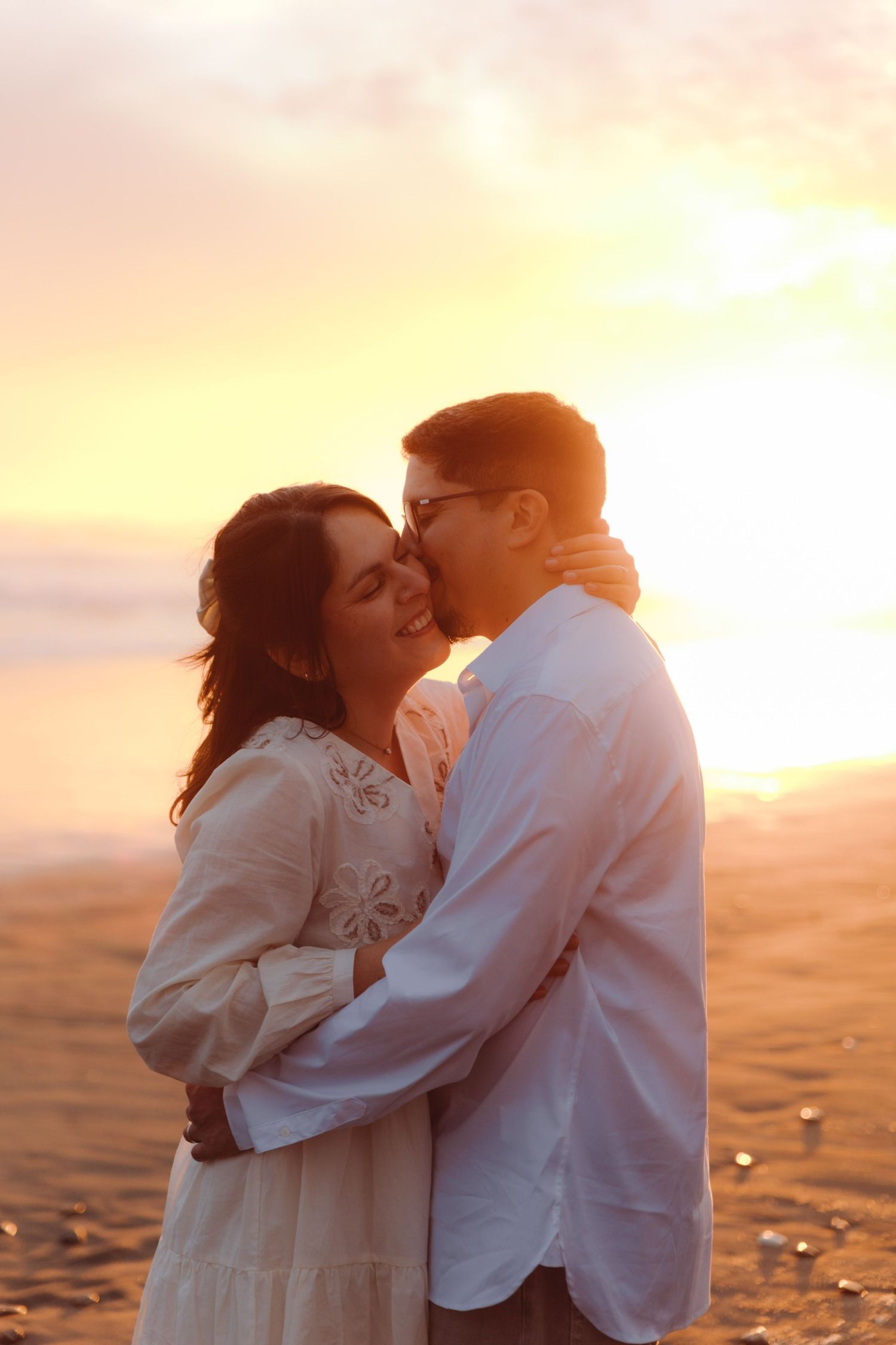 Man tenderly kissing smiling woman on cheek while embracing on sandy beach at sunset.