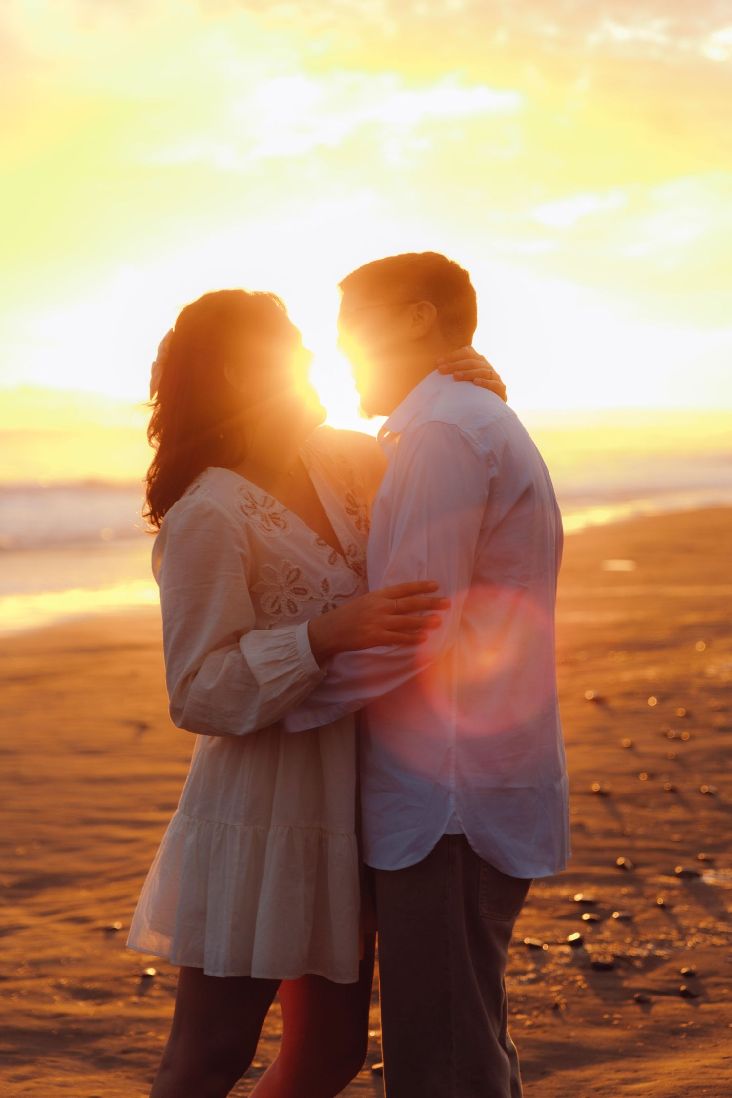 Couple holding each other close facing sunset on beach with bright golden sun behind them.