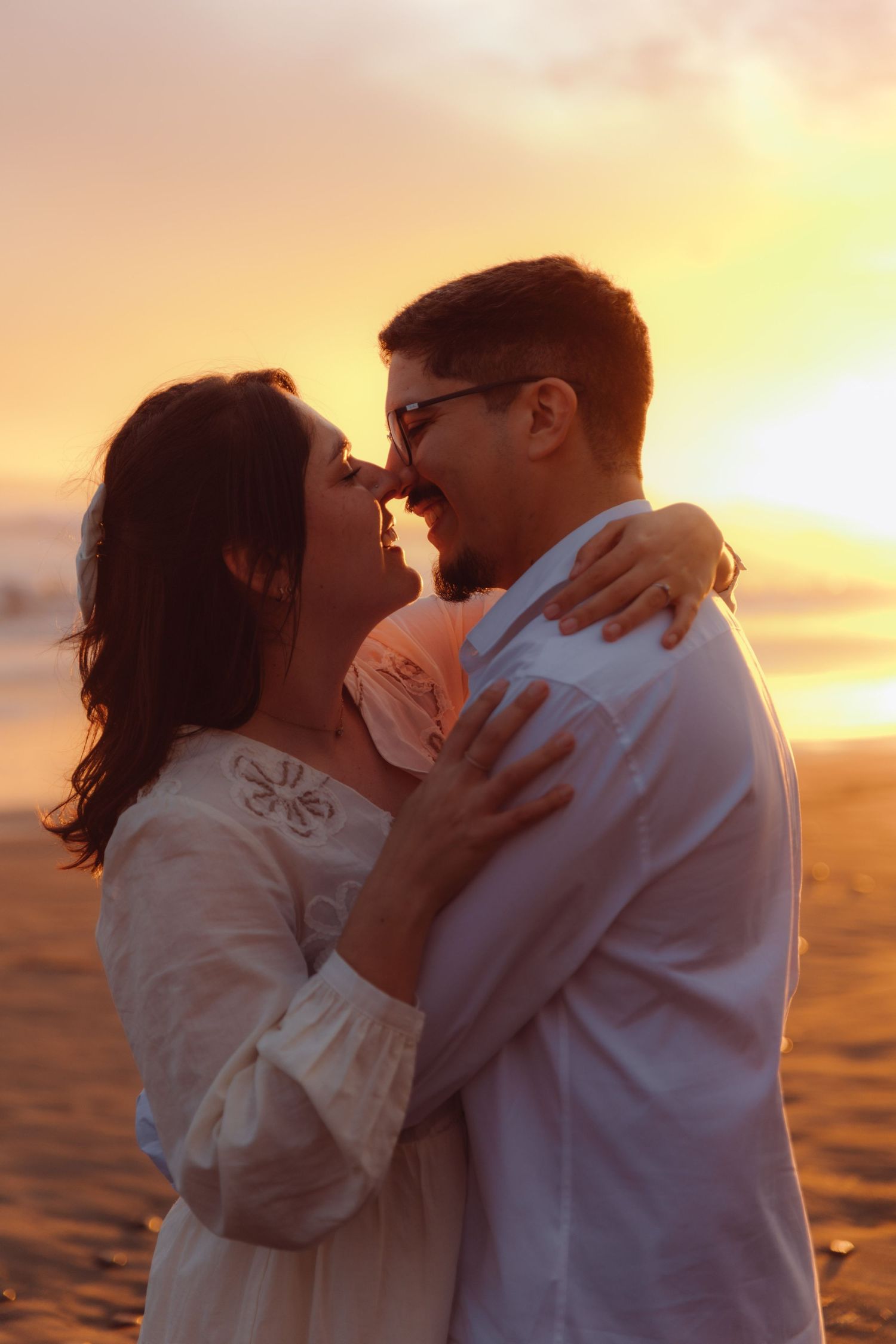 Couple about to kiss while embracing on beach during warm glowing orange sunset.