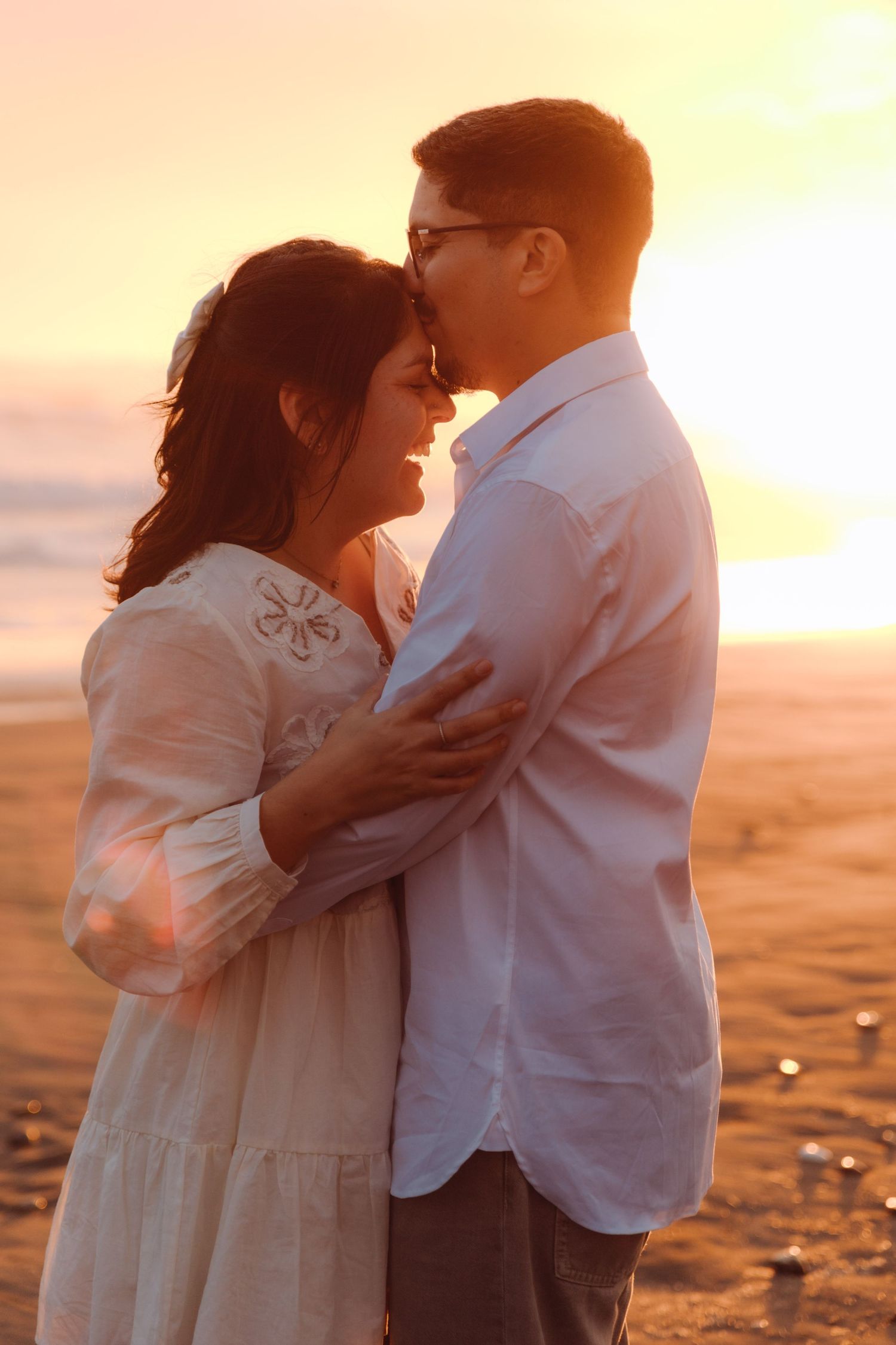 Couple holding each other close on sandy beach bathed in warm orange sunset glow.