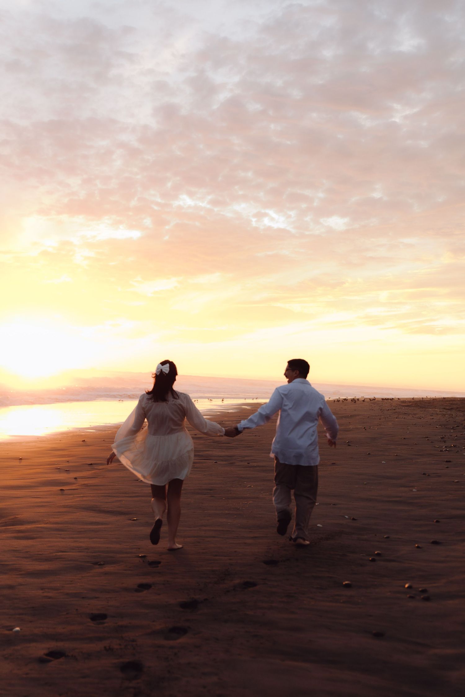 Couple holding hands running toward stunning pink and gold sunset on wide sandy beach.