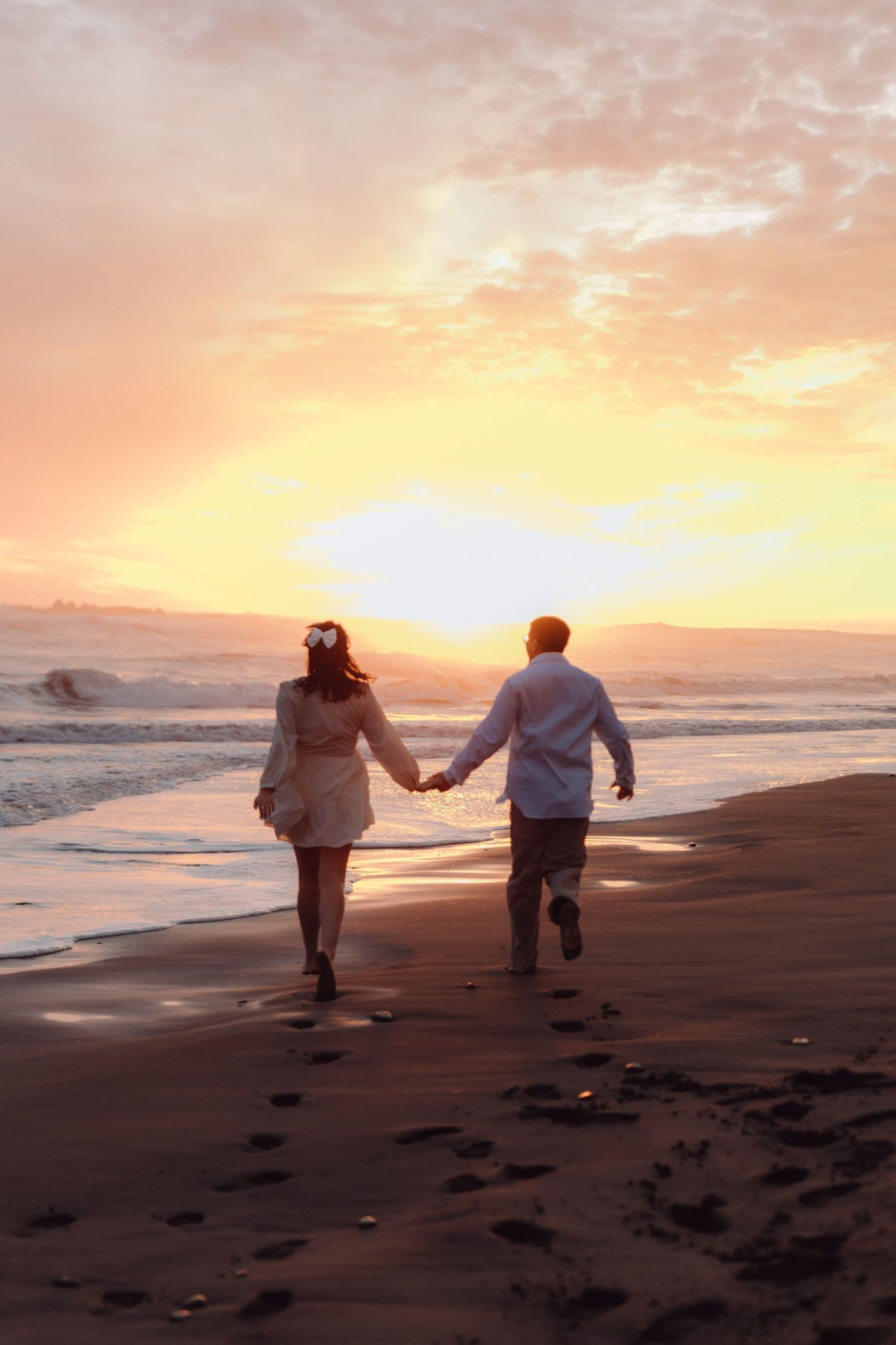 Couple running hand in hand along shoreline with vibrant golden sunset over ocean waves.
