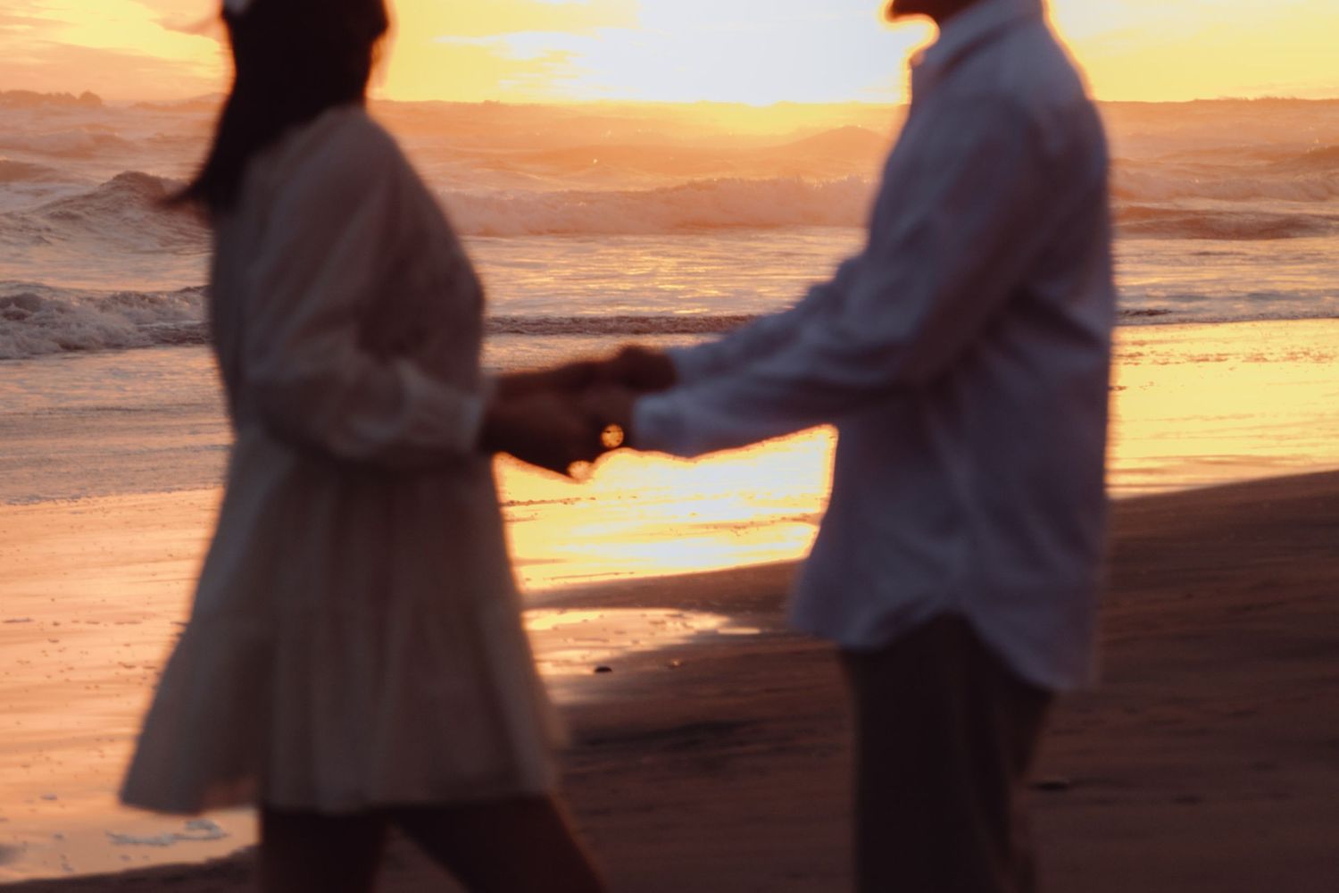 Blurred romantic couple holding hands on beach at sunset with golden light on water.