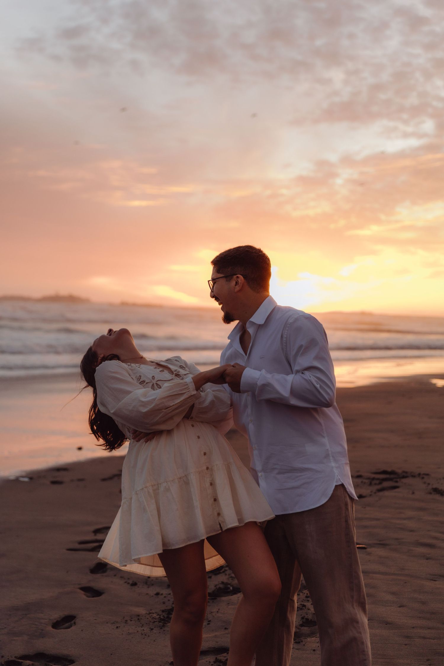 Joyful couple dancing and laughing together on beach during breathtaking pink and orange sunset.
