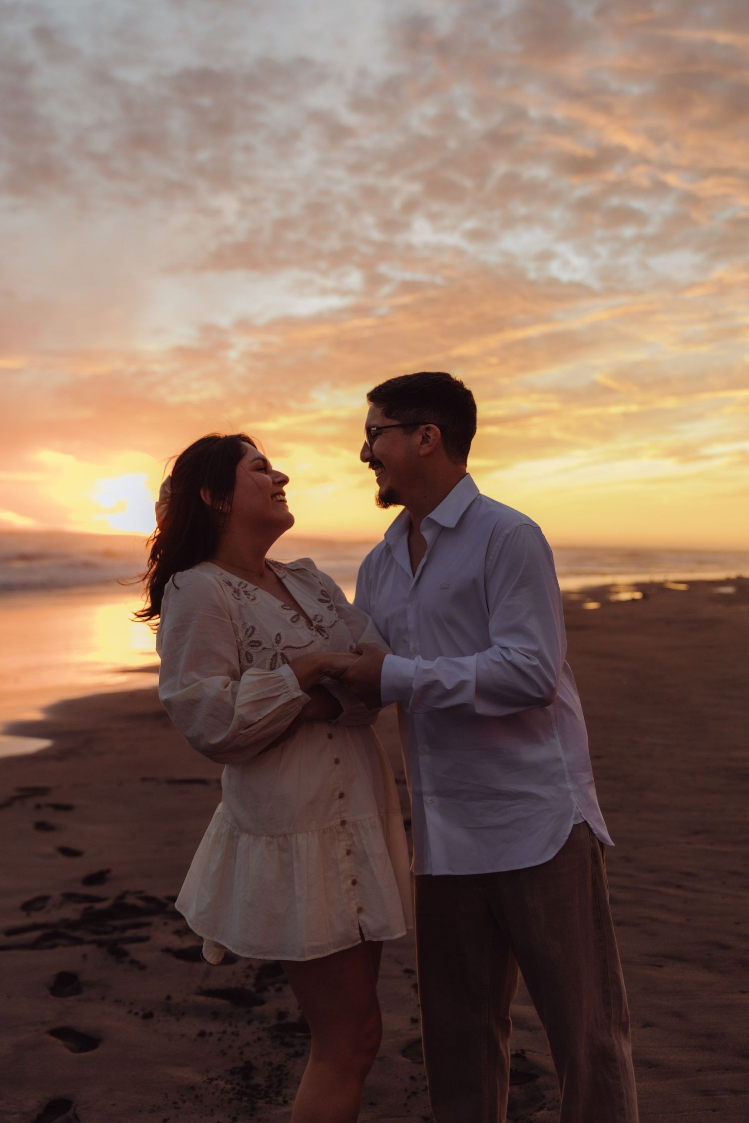 Happy couple holding hands and smiling at each other on beach at colorful sunset.