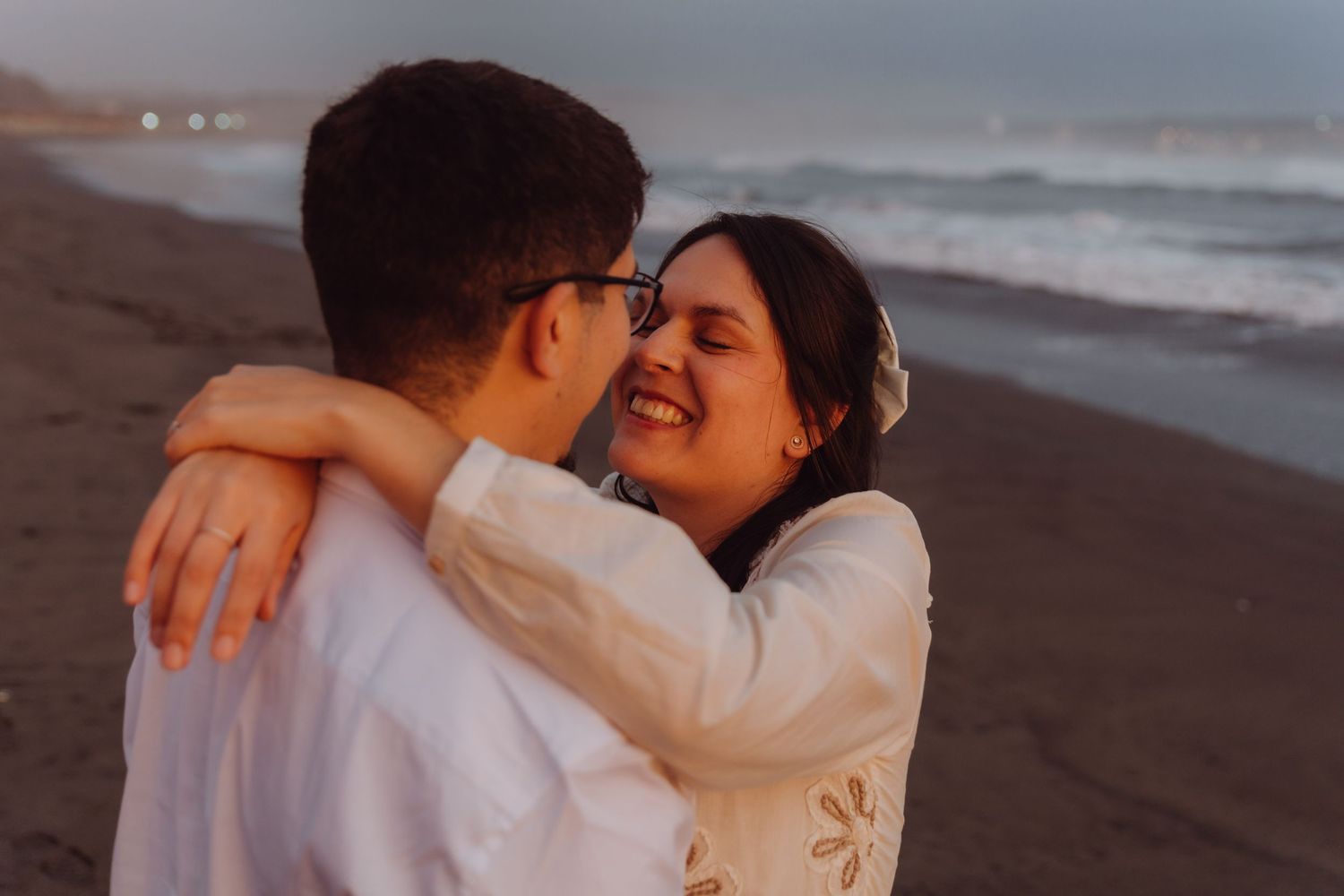 Smiling woman embracing man on sandy beach at sunset, wearing white bohemian dress.