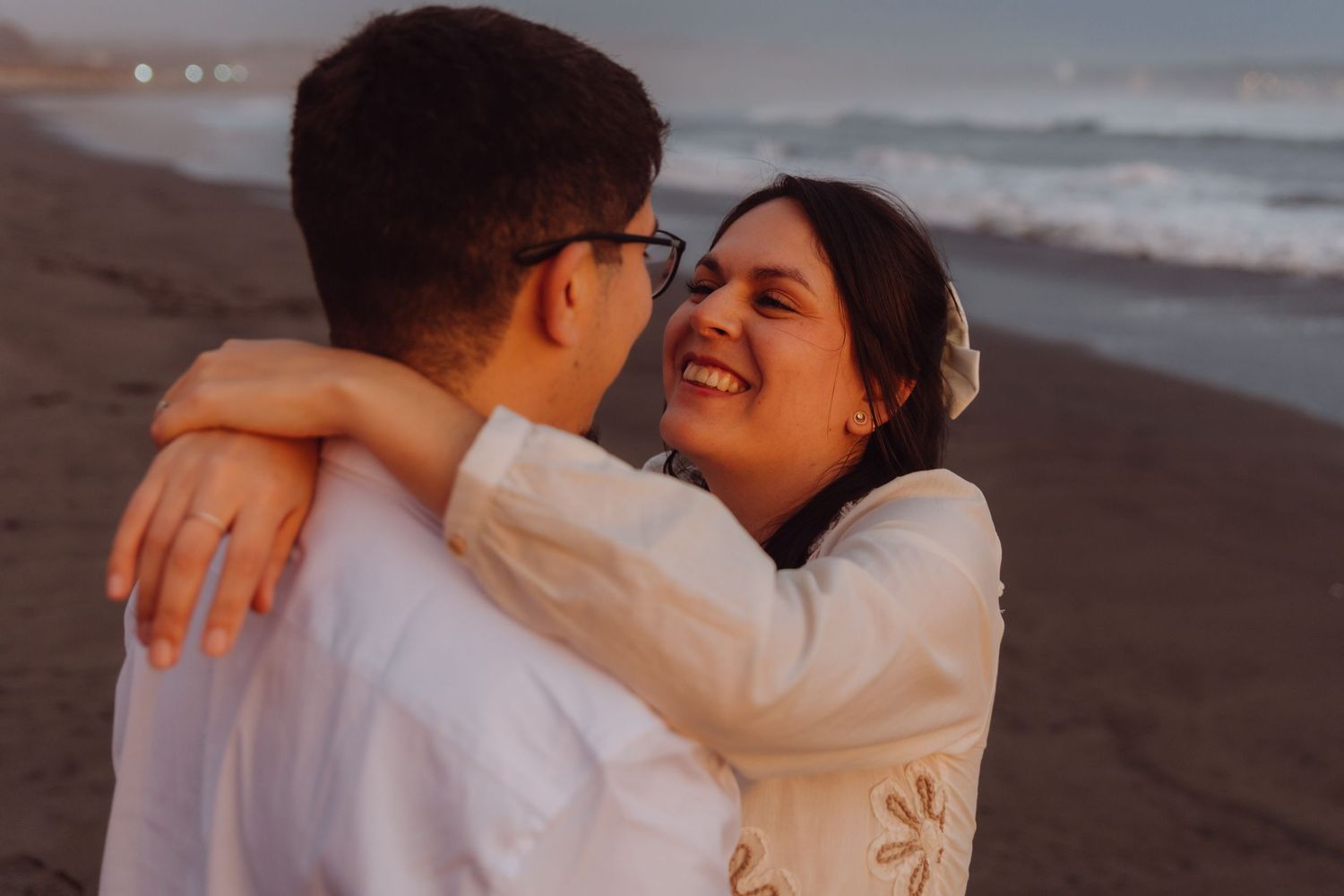 Laughing woman hugging partner on beach at golden hour, wearing white embroidered dress.