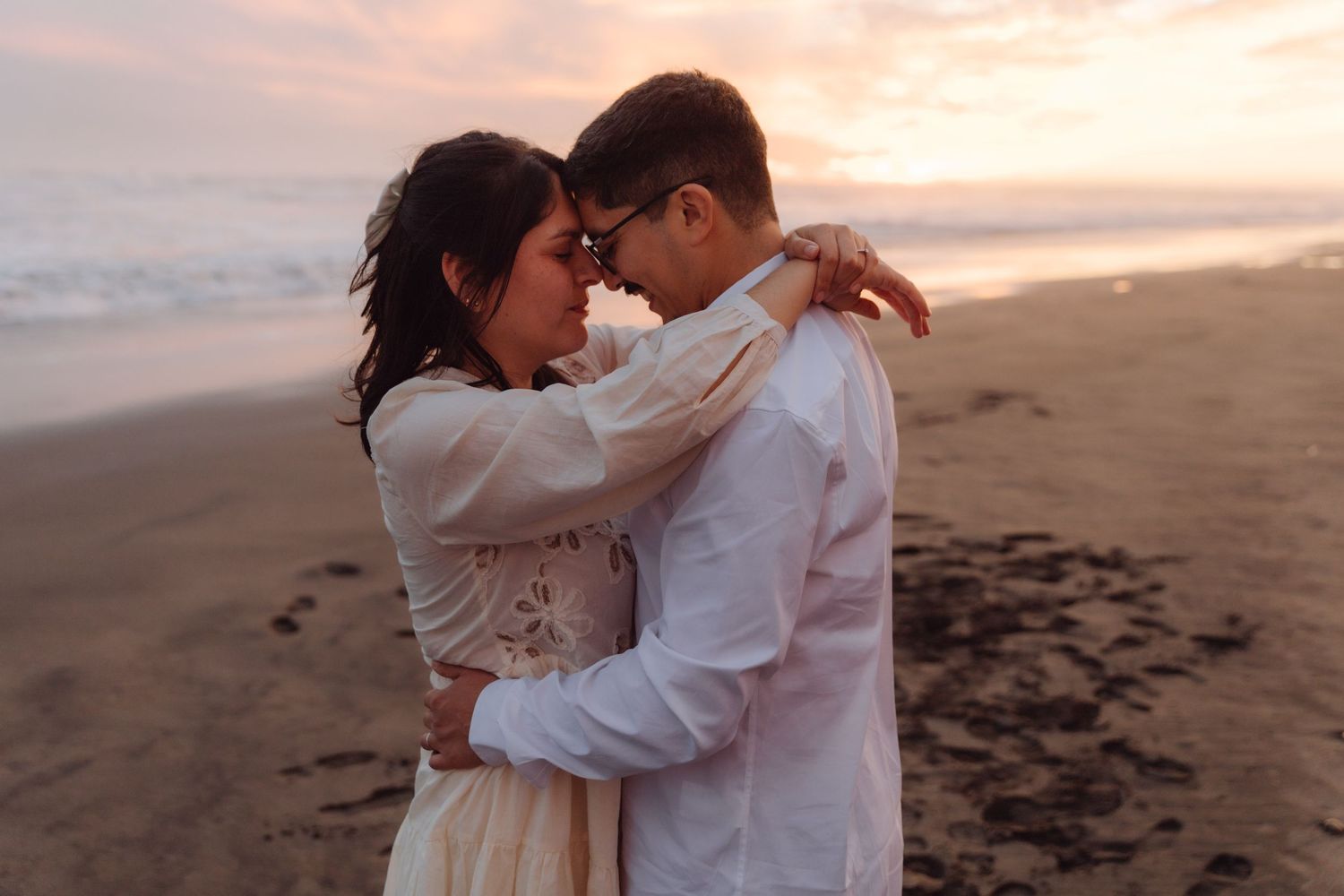 Couple tenderly touching foreheads in romantic embrace on beach during soft pastel sunset.