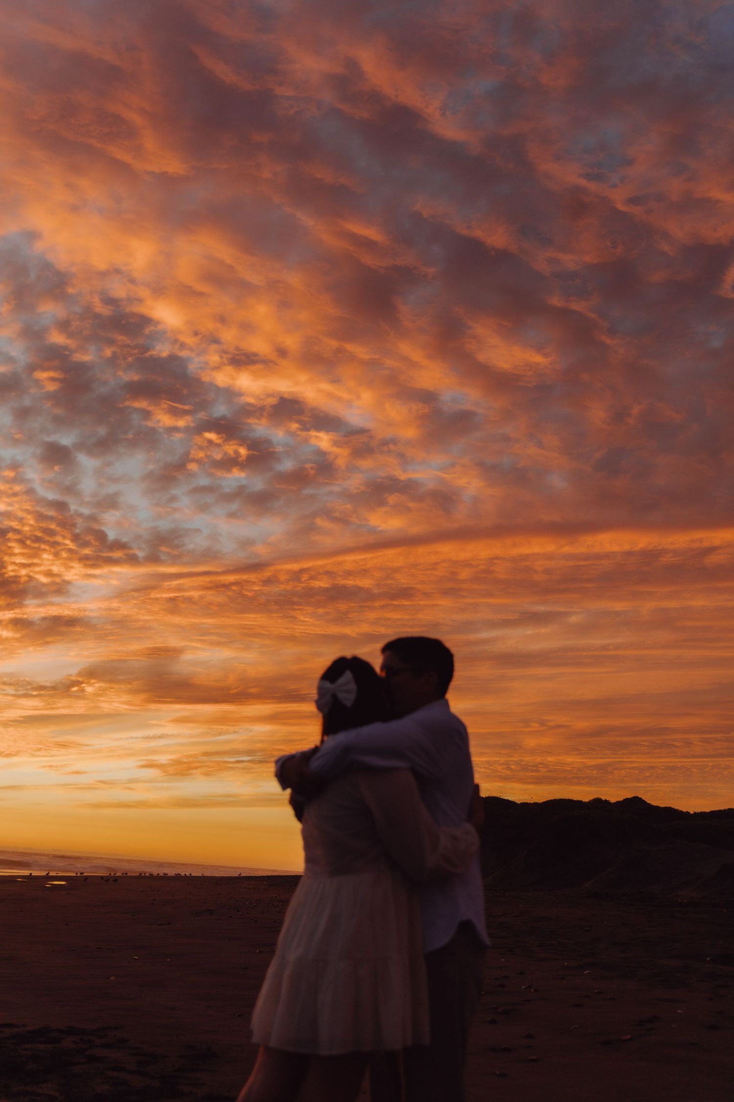 Couple hugging on beach at golden hour with dramatic orange cloudy sky.