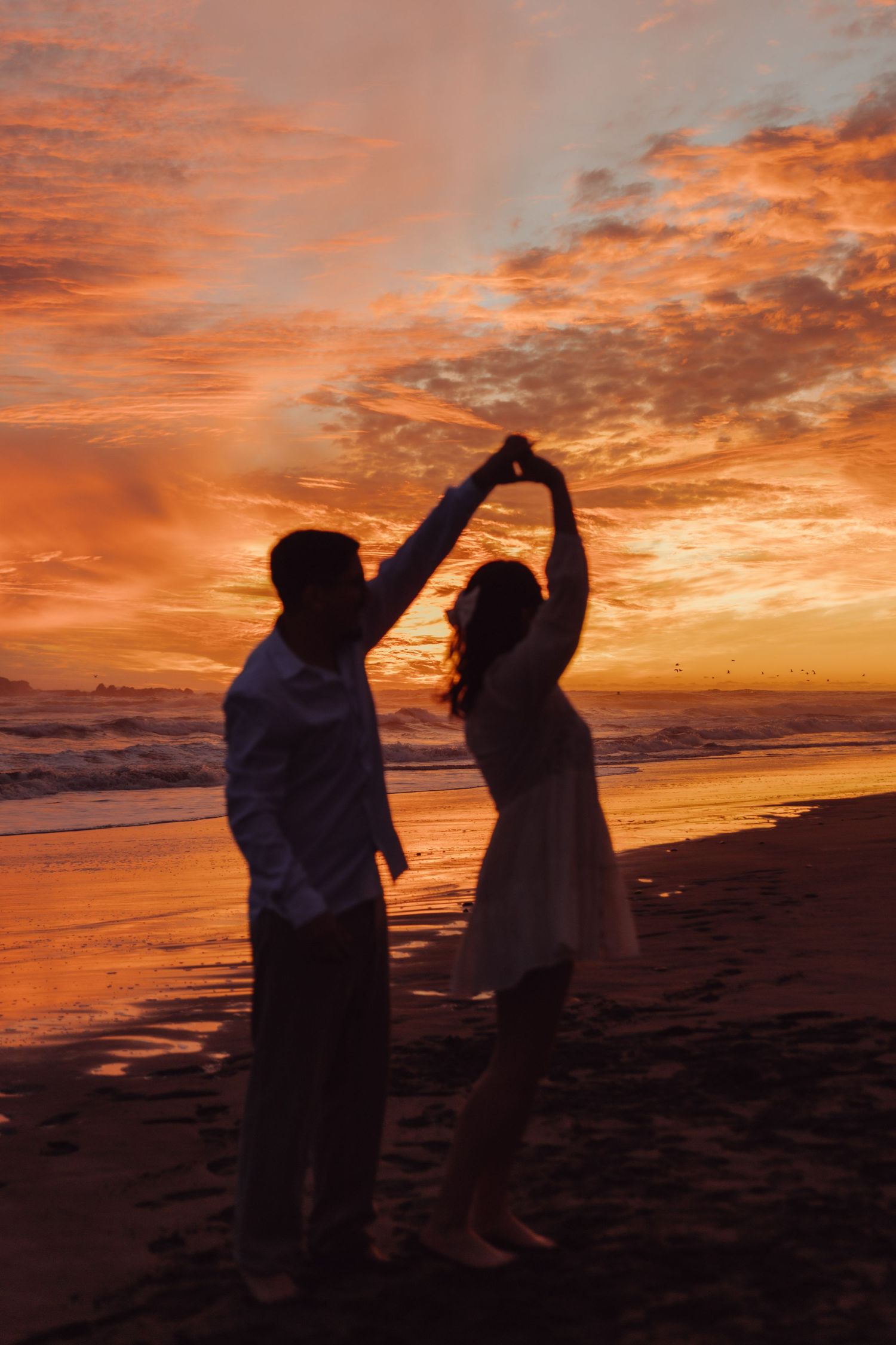 Couple dancing on beach at sunset, man spinning woman under vibrant orange sky.