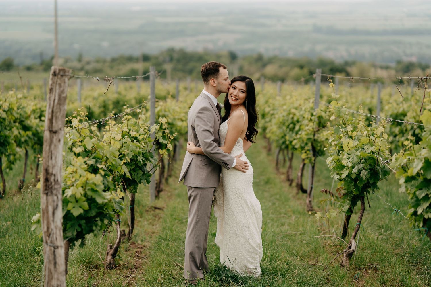 Couple embracing in a lush vineyard on their wedding day, bride in white gown and groom in tan suit.