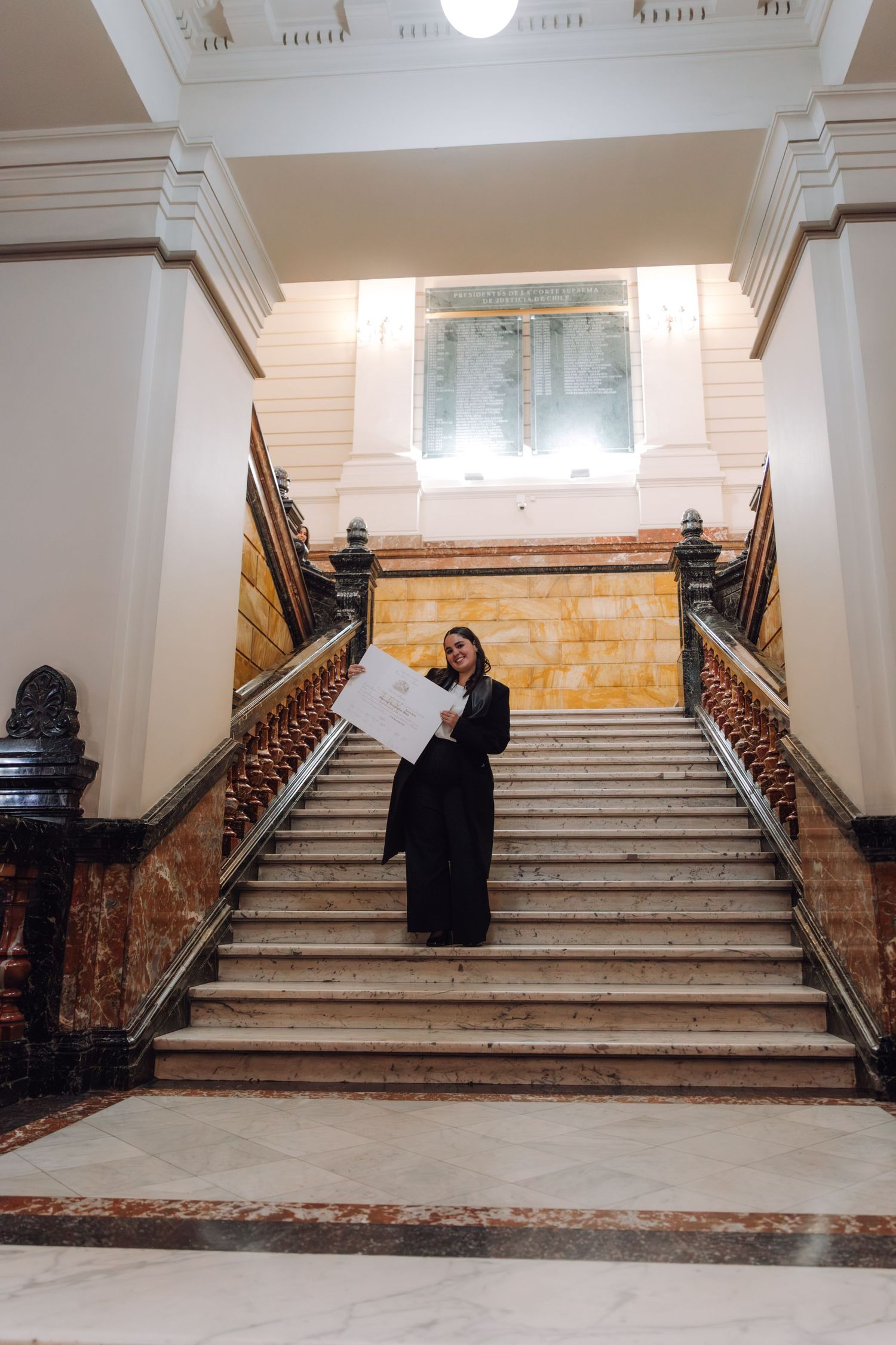 Graduate stands on grand marble staircase holding diploma in historic building.