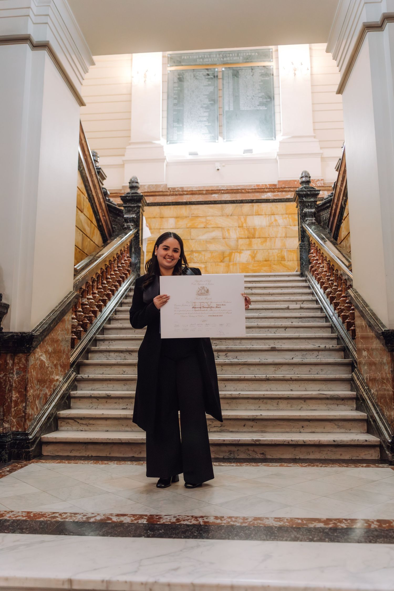 Proud graduate holds diploma at base of marble staircase in historic building.