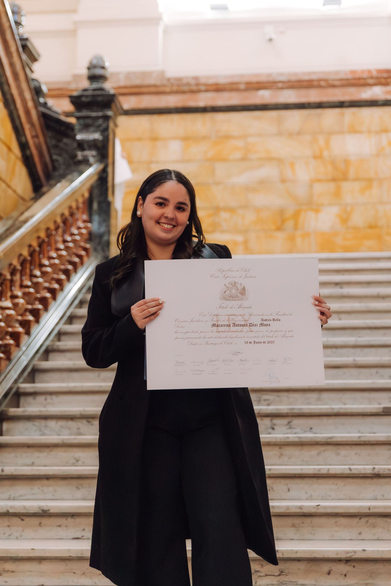 Young woman in black outfit proudly holds graduation diploma on marble staircase.