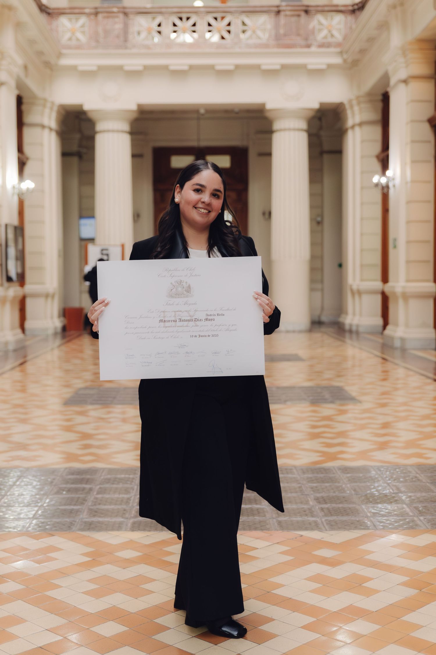 Smiling graduate in black suit displays diploma in elegant columned hall.