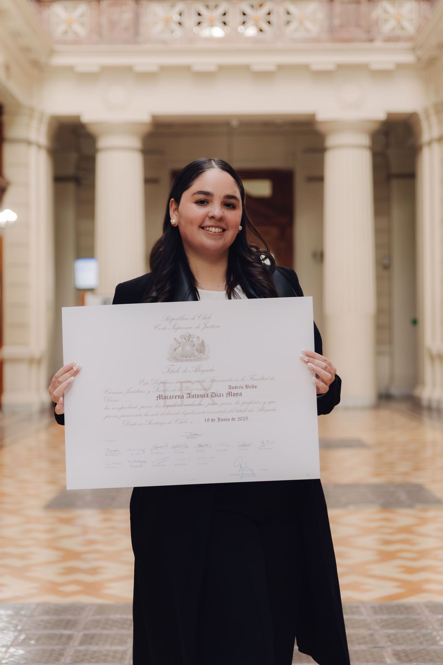 Happy young woman showcases official graduation certificate in ornate building interior.